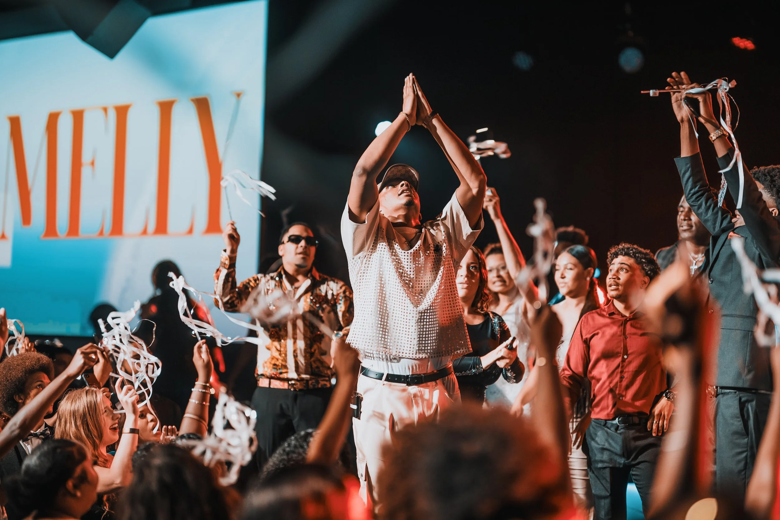 People gathered on stage at a concert or event, with some raising their hands and confetti falling, in front of a large screen displaying the word 'NIELY' in bold letters.