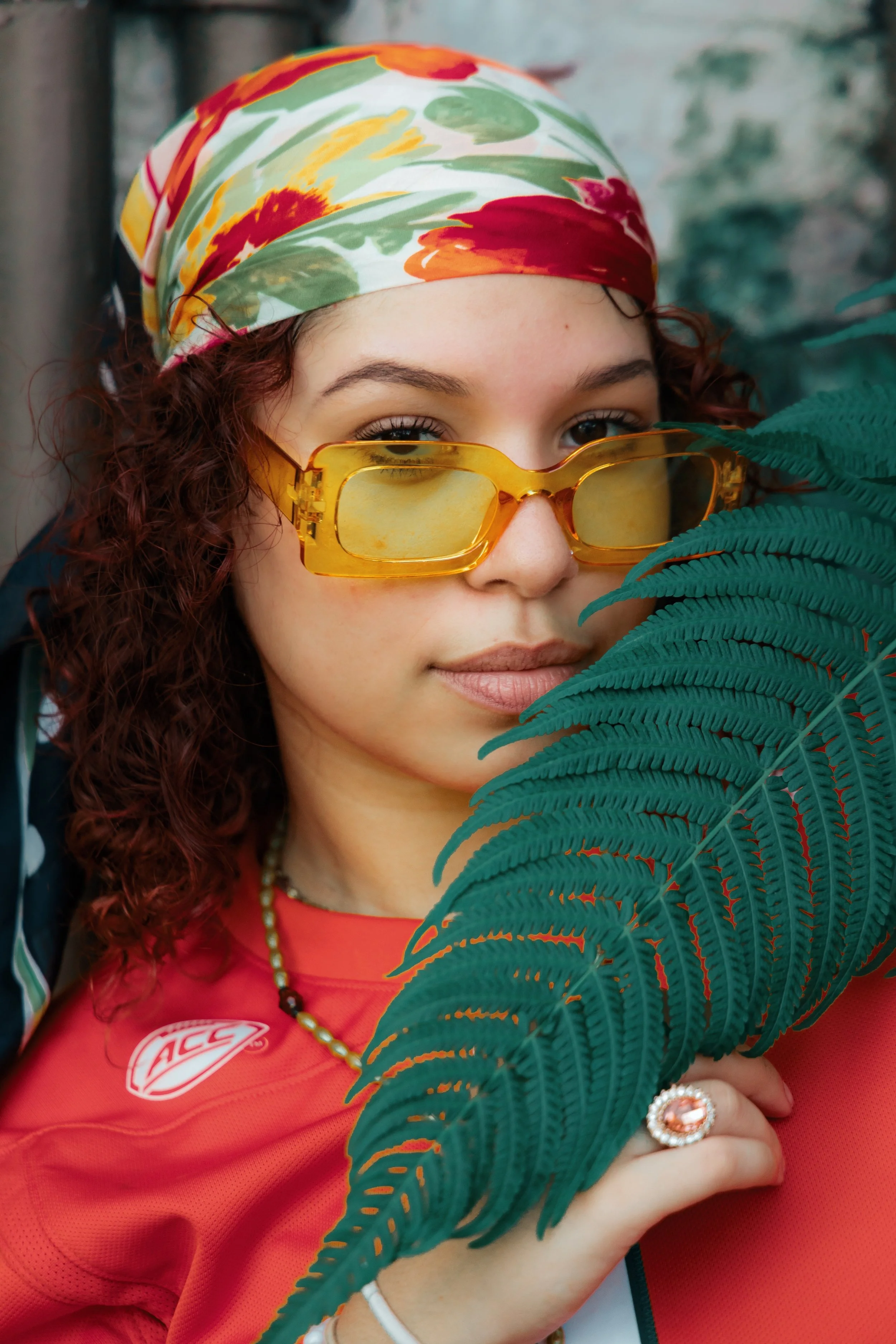 A young woman with curly red hair wearing a colorful headscarf, yellow sunglasses, and a coral-colored sports jersey, holding a large fern leaf in front of her face.