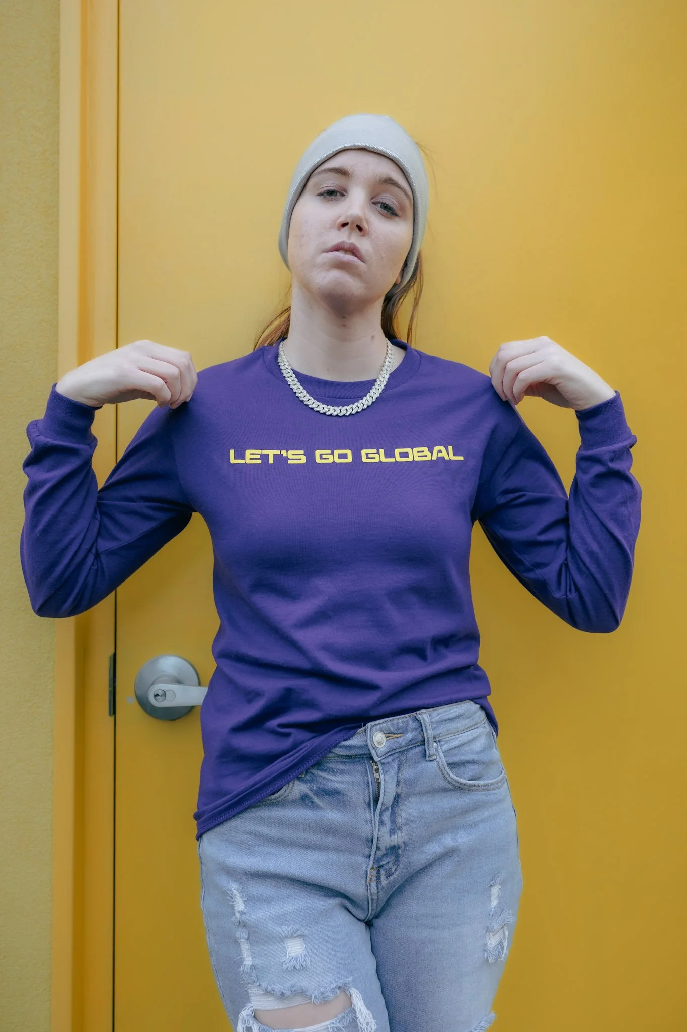 A woman with a serious expression, wearing a white beanie, a purple sweatshirt with yellow text that says 'Let's Go Global,' gold chain necklace, and ripped jeans, standing against a yellow door background.