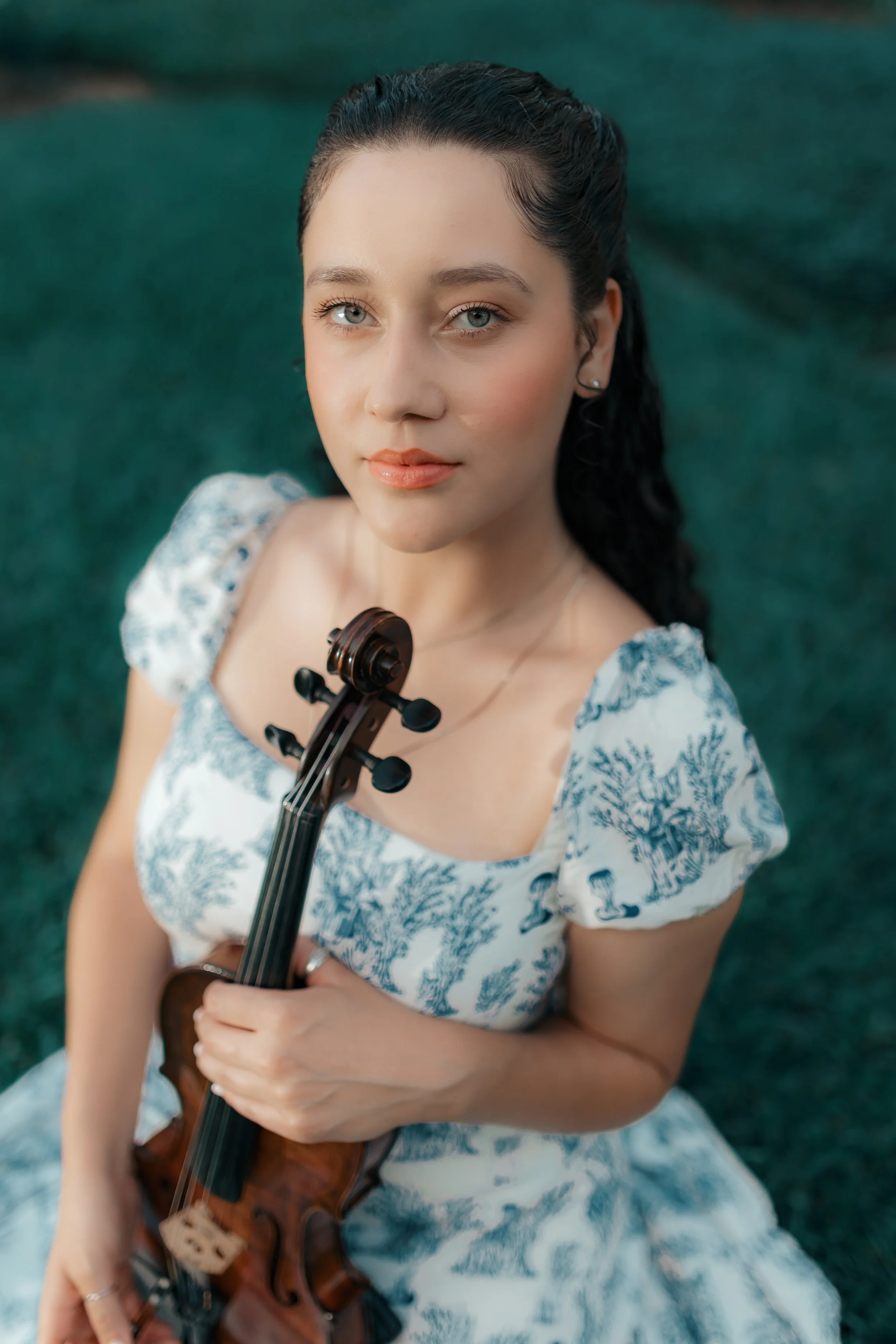 A young woman with long dark hair and light skin, wearing a blue and white patterned dress, holds a violin and looks directly at the camera outdoors on a grassy area.