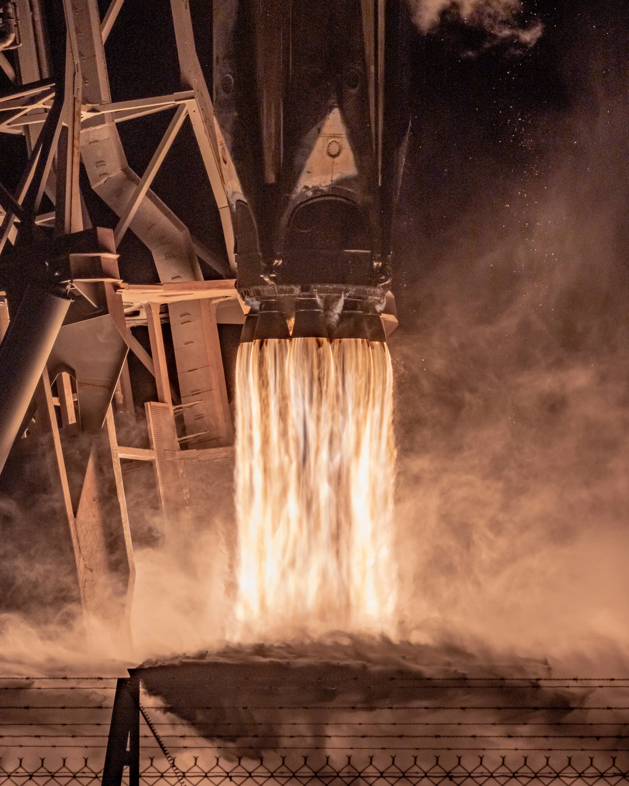 A rocket booster is igniting during launch, emitting a large bright flame and exhaust fumes into the dark sky.
