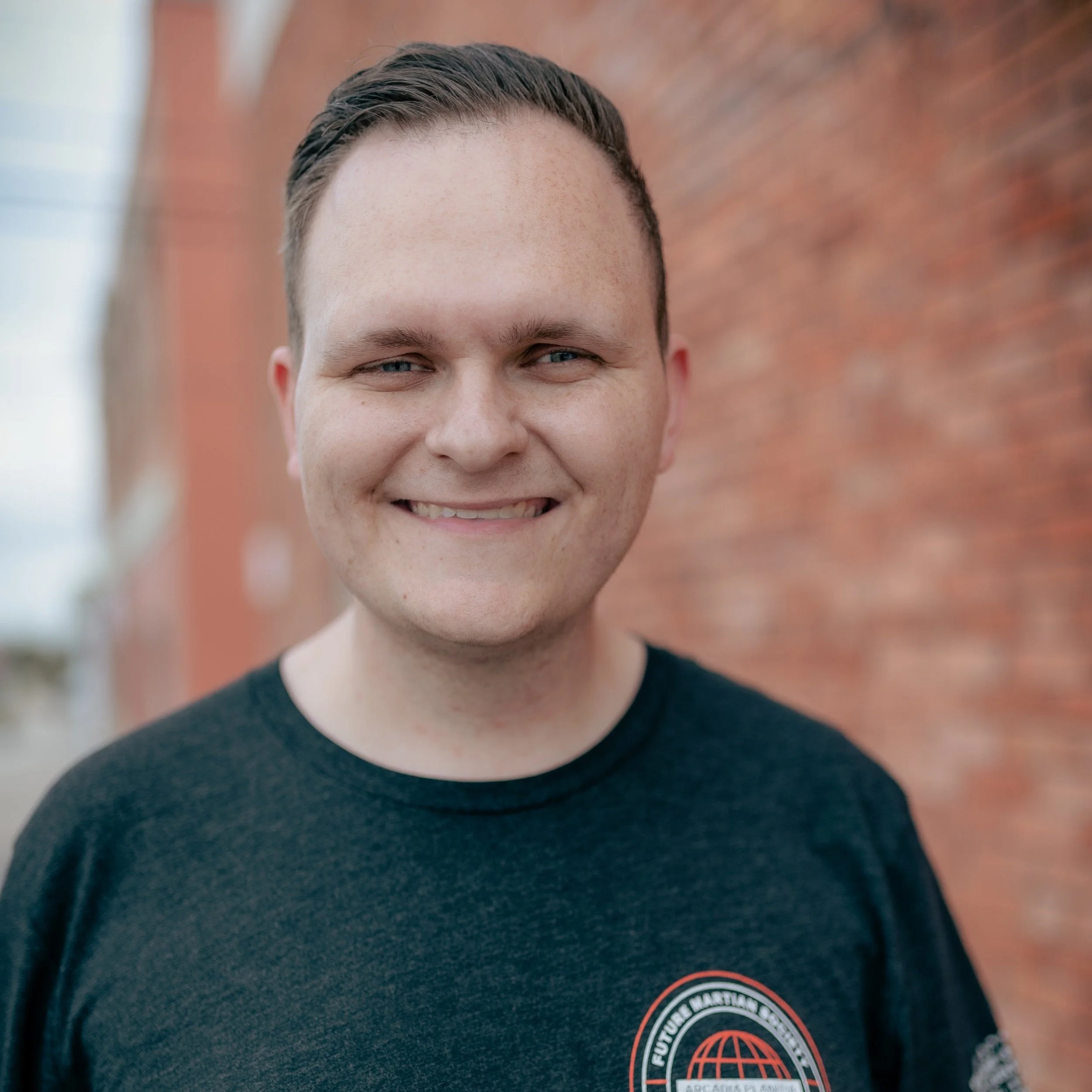 Smiling young man with short brown hair wearing a black t-shirt with a logo, standing outdoors near a red brick wall.