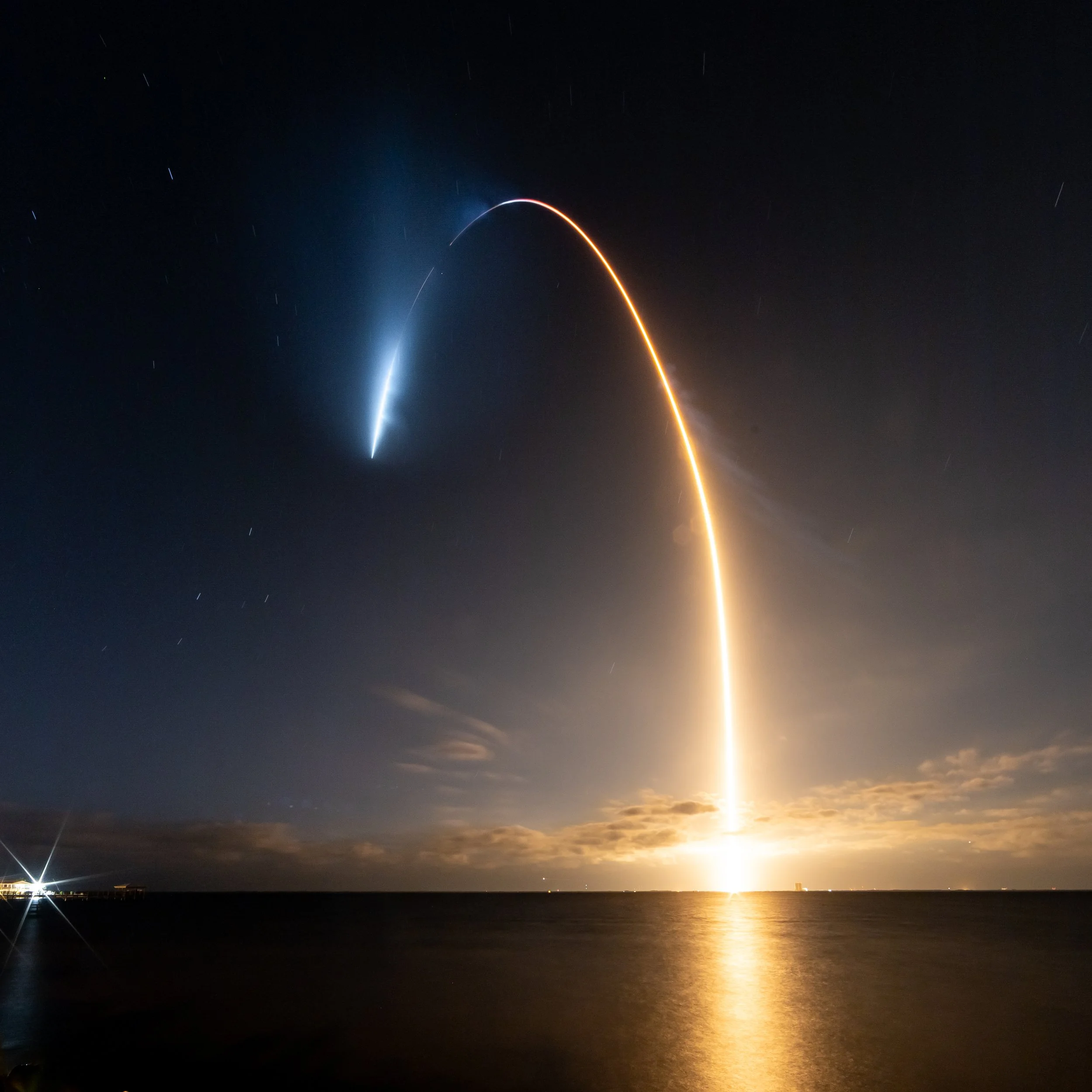 A rocket launching at night, with a bright fiery trail reflecting on the water, and a streak of smoke and fire trailing upward into the sky.
