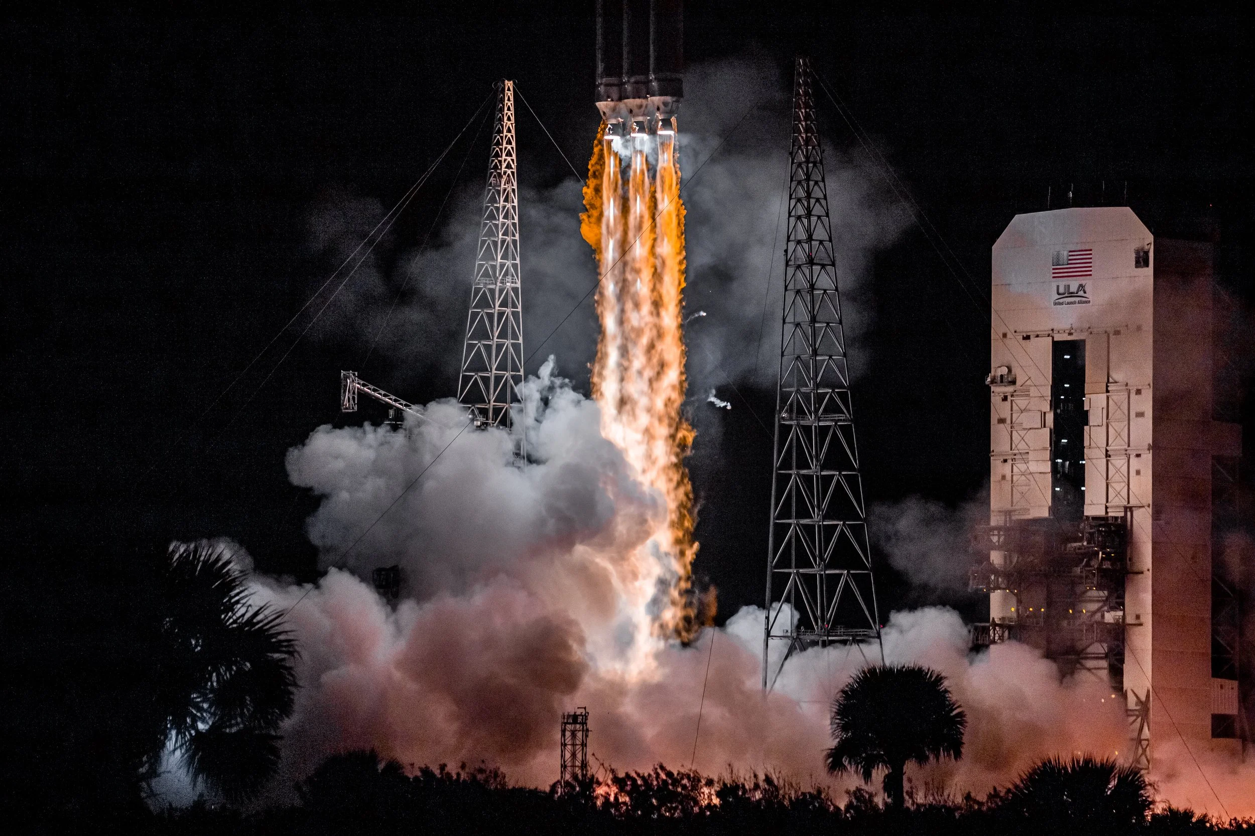 A rocket launch at night with flames and smoke coming from the rocket engine, surrounded by launch tower structures and palm trees in the foreground.