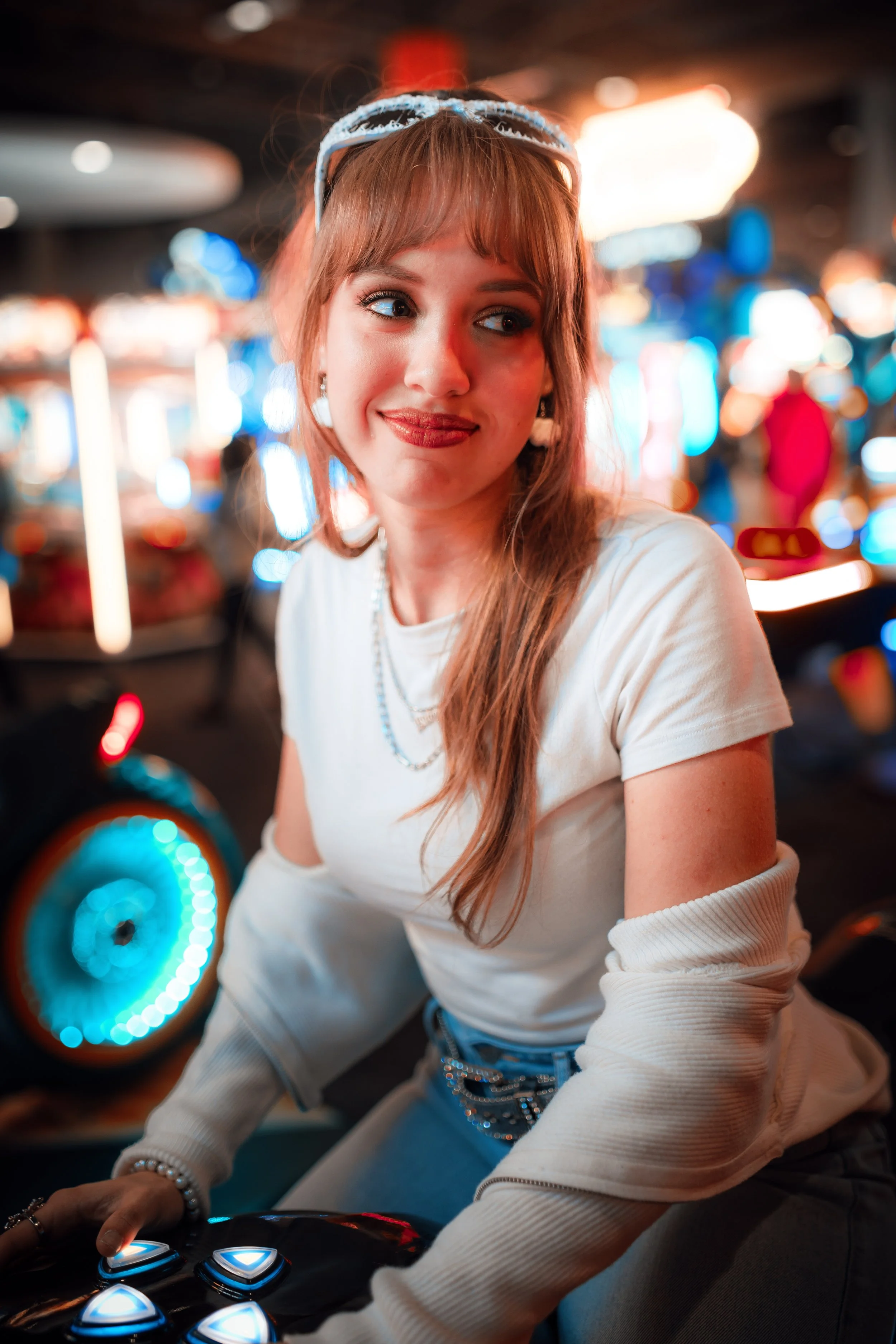 A young woman with red hair and fair skin in a white shirt is in an arcade, surrounded by colorful lights. She wears a headband, earrings, layered necklaces, and a bracelet, and she has a playful, smiling expression as she looks to the side.