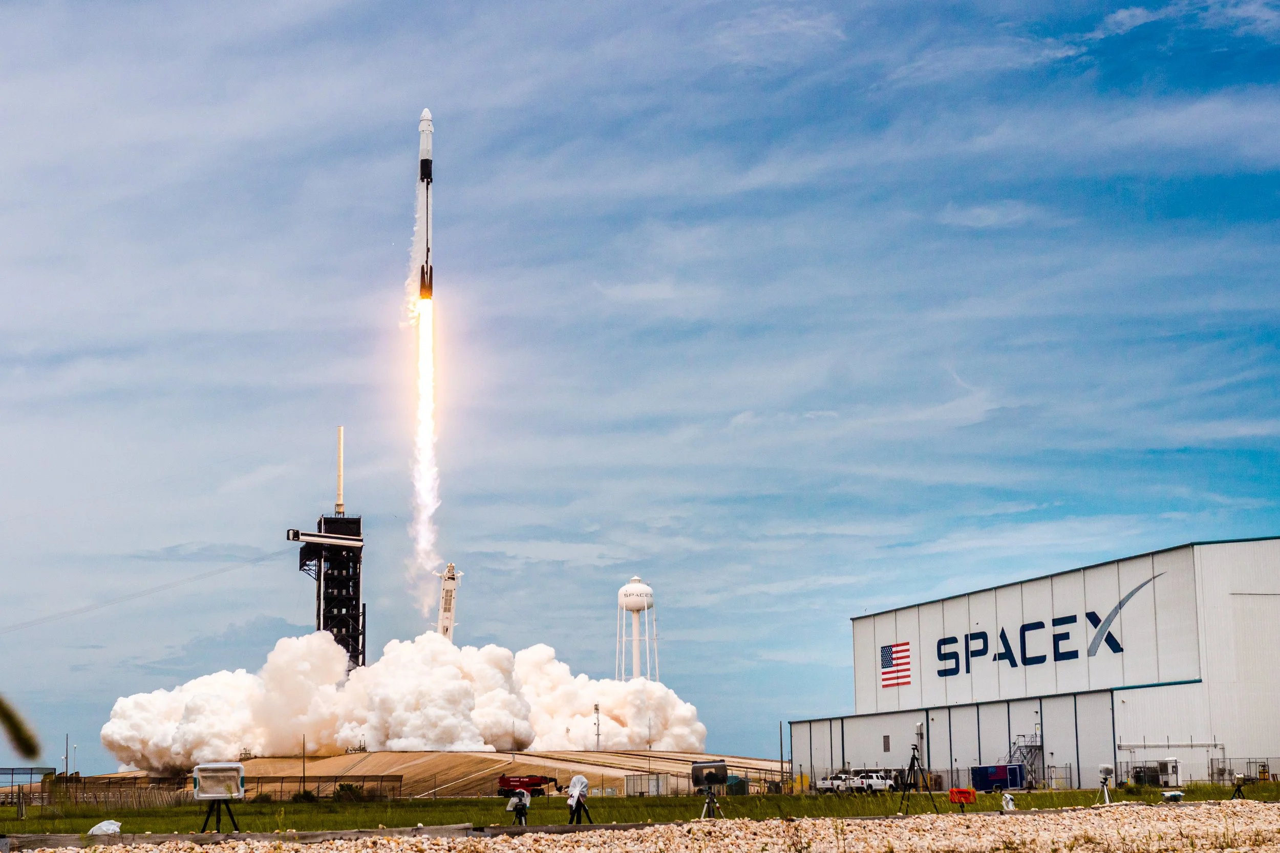 A SpaceX rocket launching into the sky with smoke and fire at launch pad, with a large SpaceX building and water tower nearby.