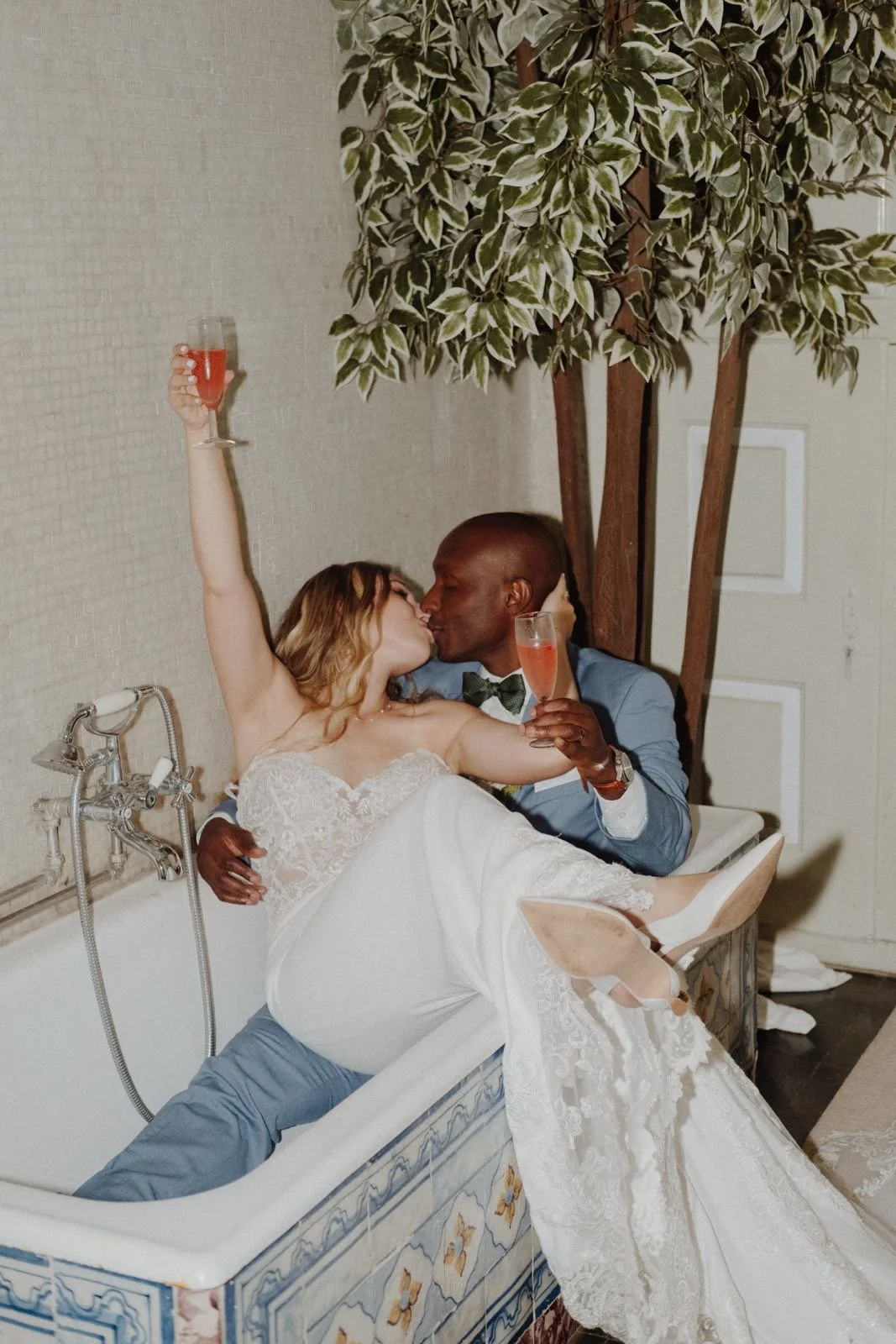 A bride and groom sharing a kiss in a bathtub, holding glasses of pink champagne, with a few trees in the background.