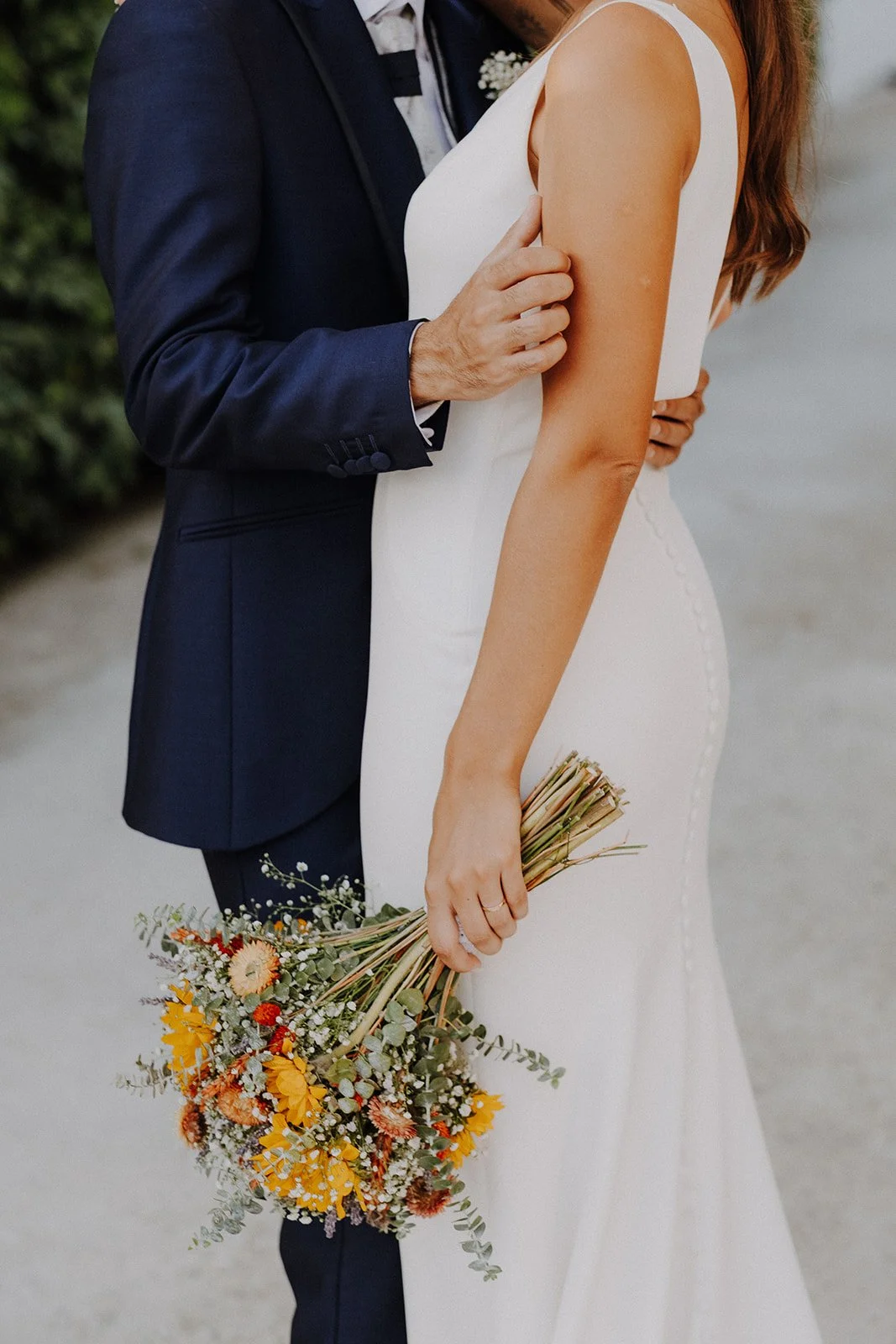 A bride in a white wedding dress holding a colorful bouquet of flowers. A groom in a dark suit is touching her arm.