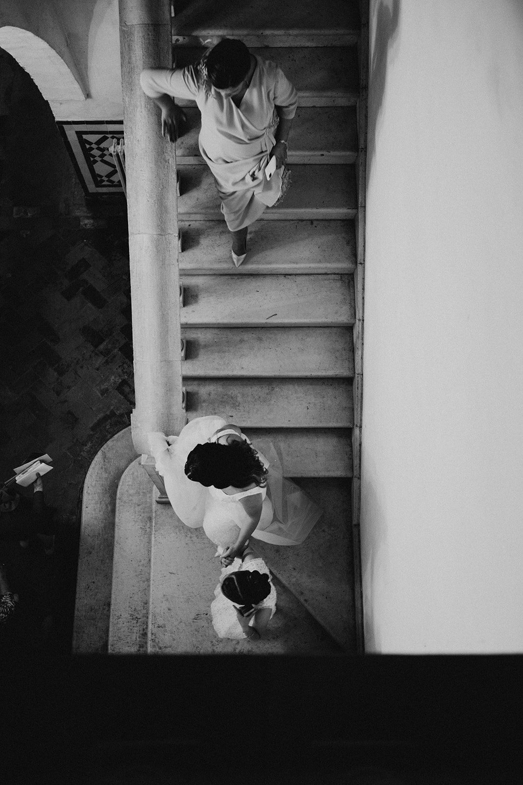A black and white photo of two women holding hands and walking up a staircase, taken from above. One woman is wearing a white dress with her hair down, and the other is wearing a light-colored dress with curly hair.