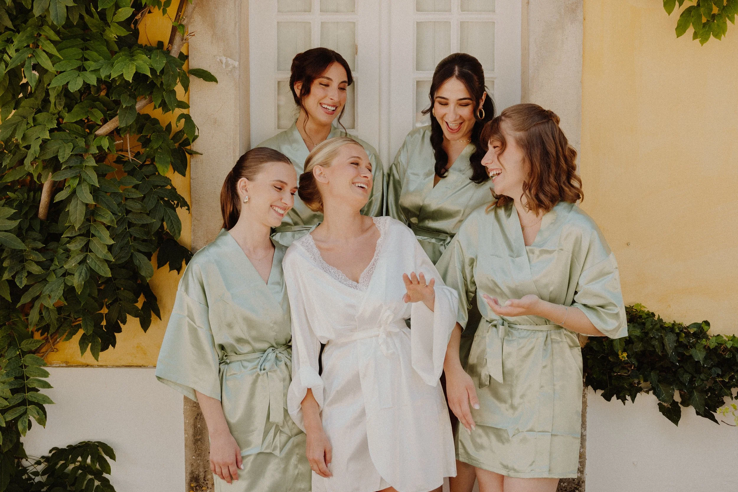 A bride and five women, possibly her bridesmaids, laughing and sharing a joyful moment outdoors, wearing matching satin robes.