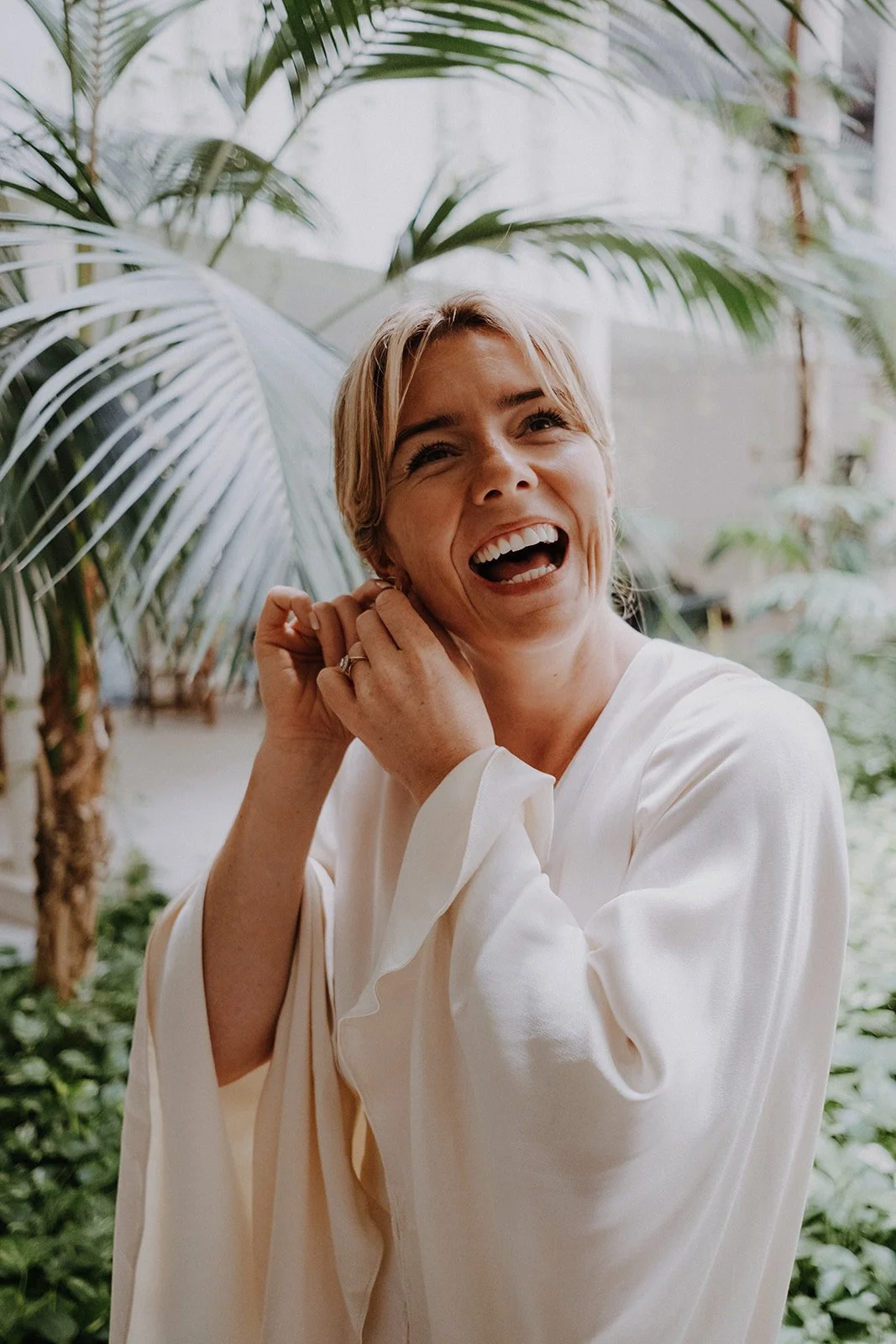 A woman laughing and adjusting her earring, standing outdoors among tropical plants.