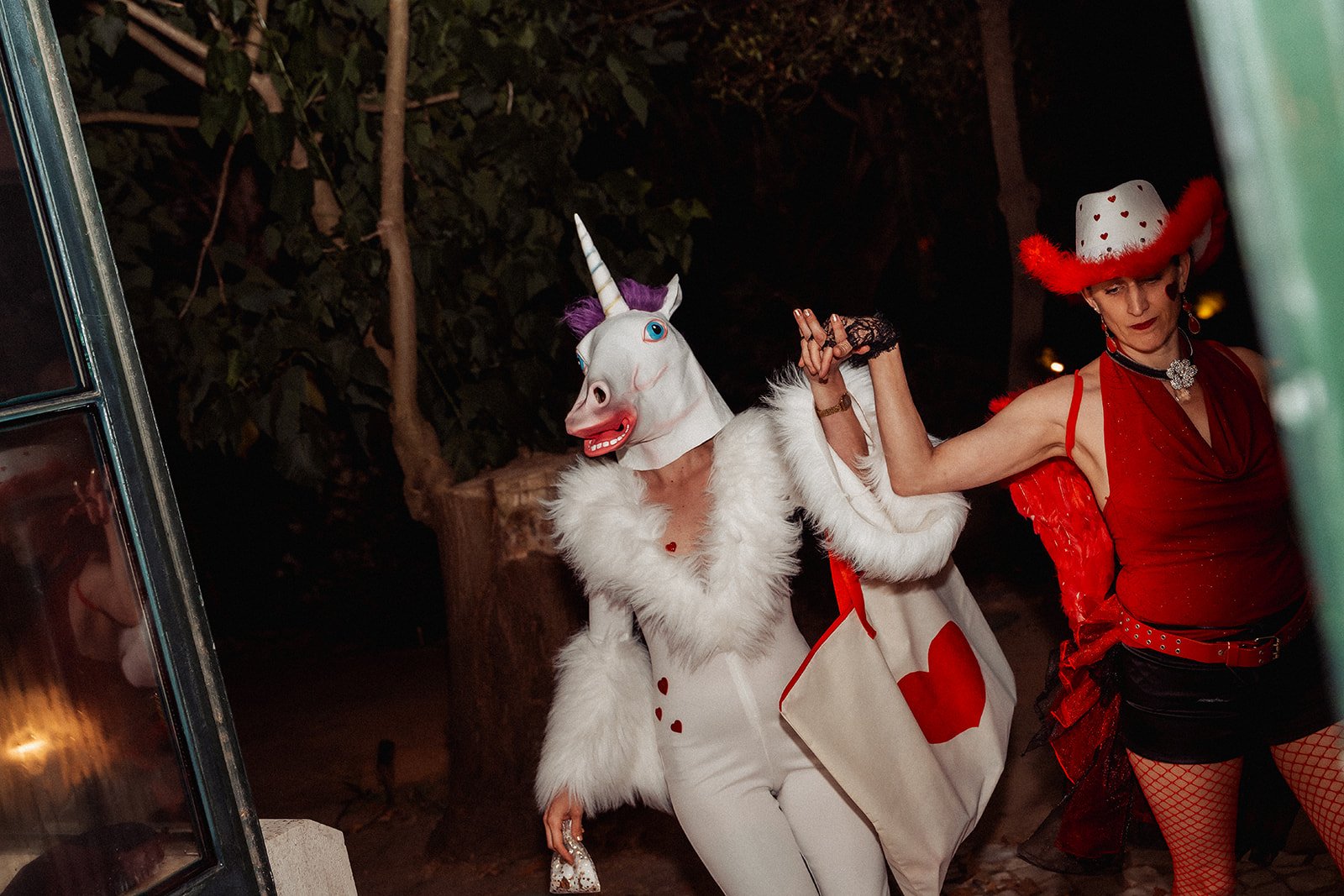 Two women dressed in costumes, one as a unicorn with a mask and the other as a woman in a red dress and large red and white hat, holding hands and dancing outdoors at night.