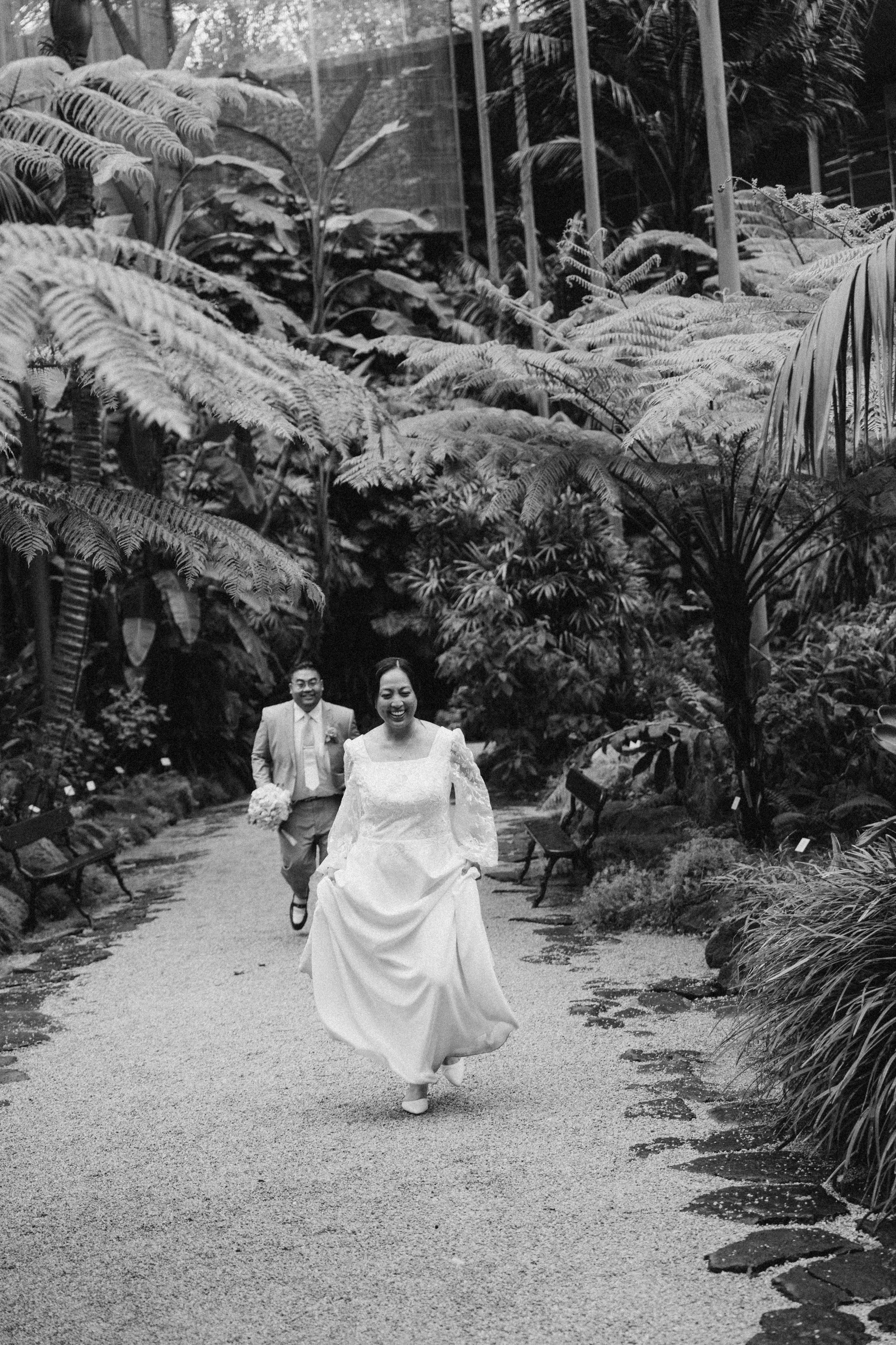 A black and white photo of a bride in a wedding dress running down a garden path with a man in a suit following her, surrounded by lush tropical plants.