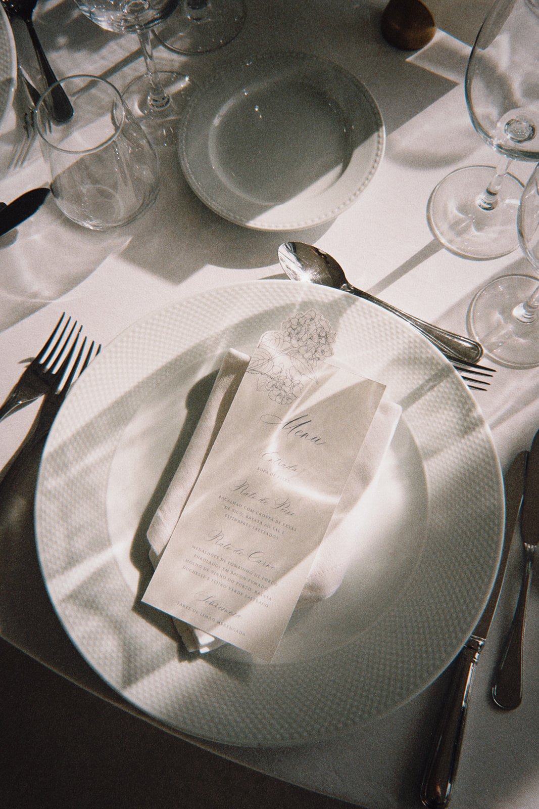 Elegant dining table setting with a white plate, silverware, wine glasses, a small bowl, and a printed menu on a napkin.