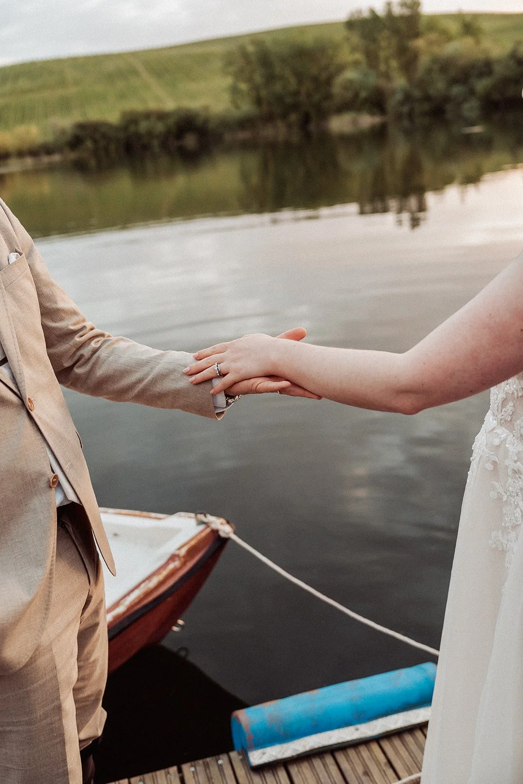 A close-up of a bride and groom holding hands on a boat docked by a river, with water and green hills in the background, during their wedding or commitment ceremony.