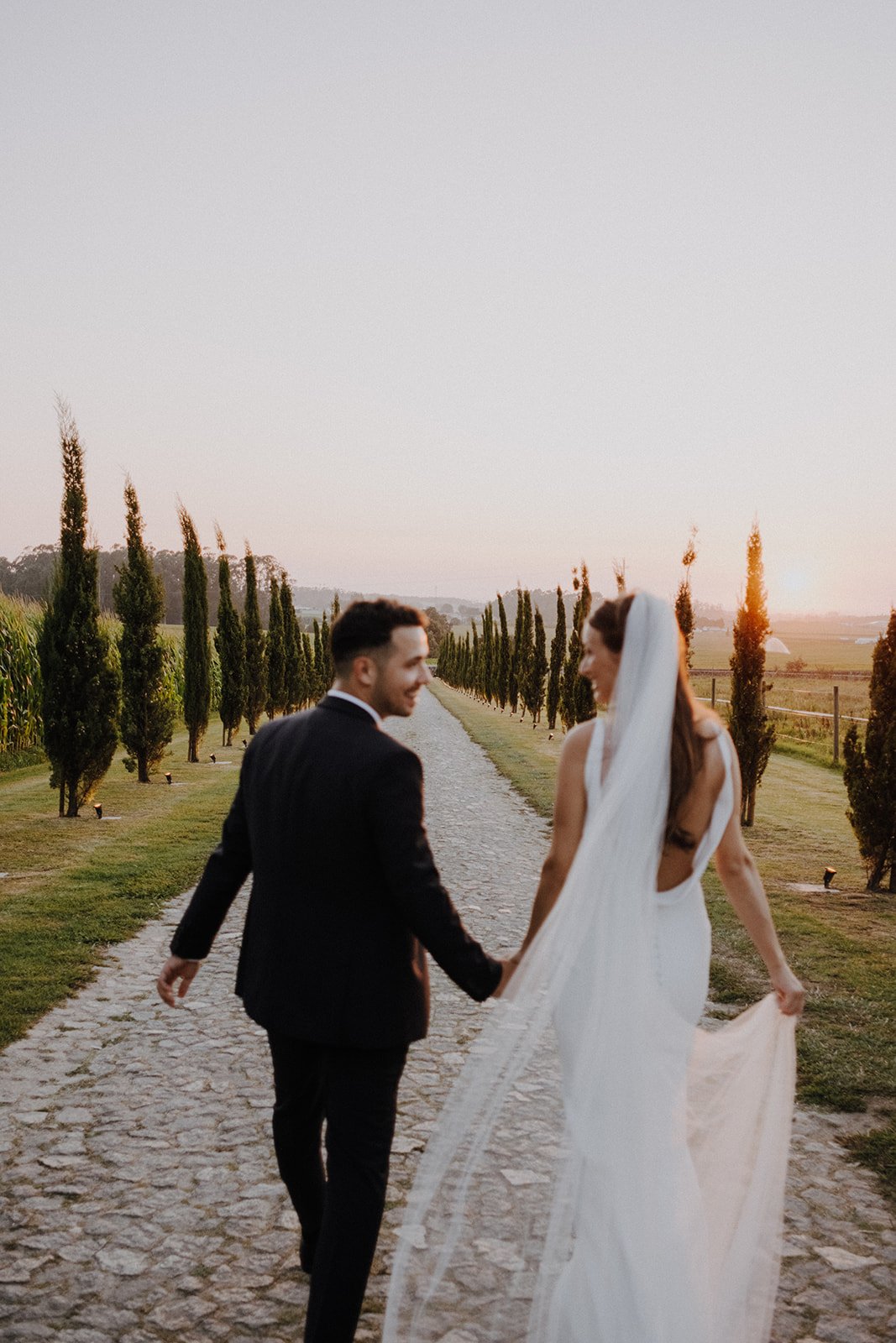 A bride and groom walking hand in hand on a stone pathway lined with tall, slender trees, during sunset.