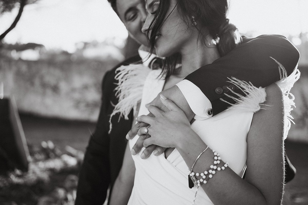 A black and white photo of a couple embracing, smiling, with the woman showing her wedding ring. They are outdoors, with a blurry background.