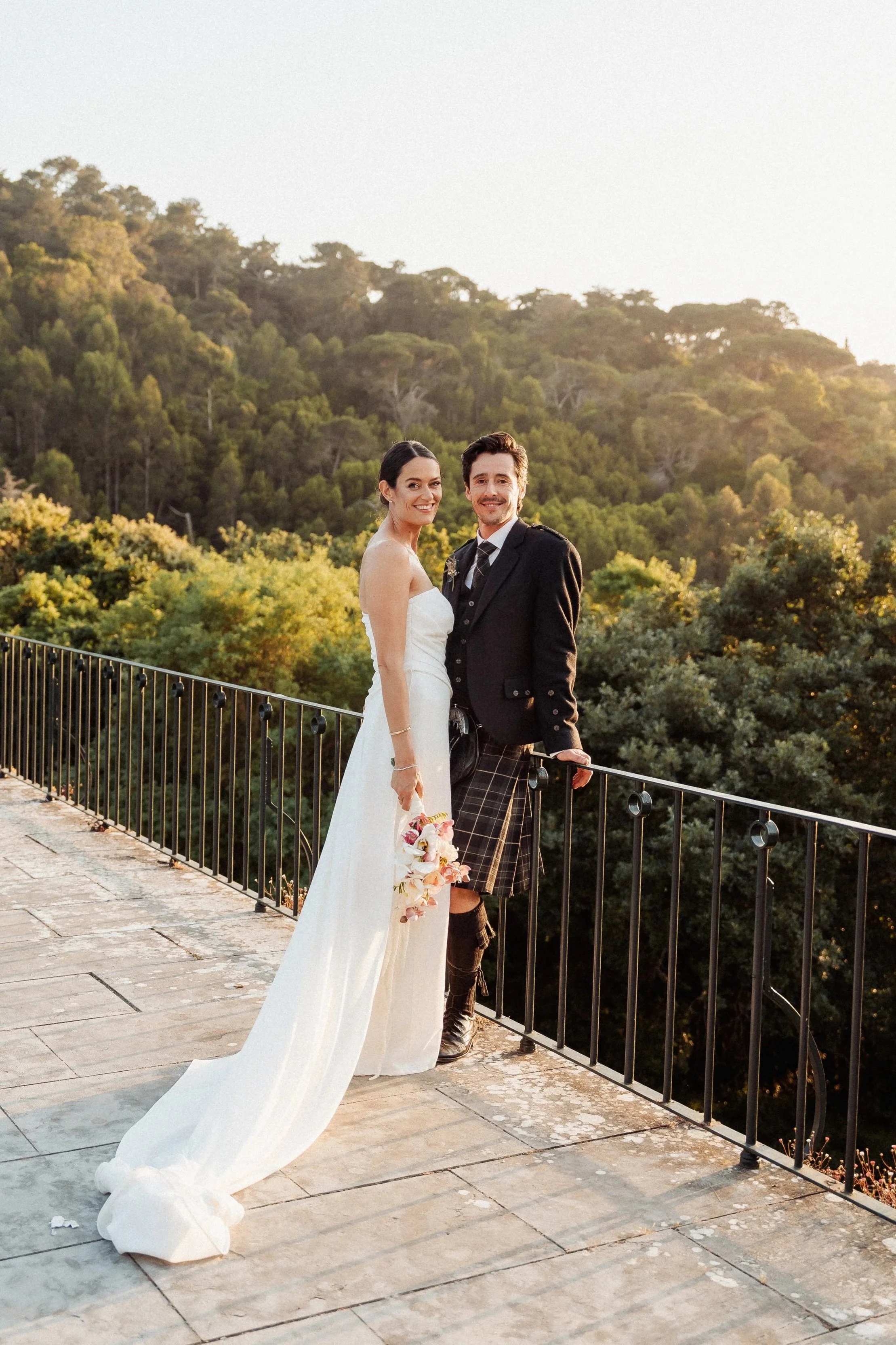A newlywed couple stands on a stone balcony with a black metal railing, surrounded by lush green trees and a hillside, during sunset. The woman is wearing a white wedding dress and holding a bouquet, while the man is in a black suit with a tartan kilt, smiling at the camera.