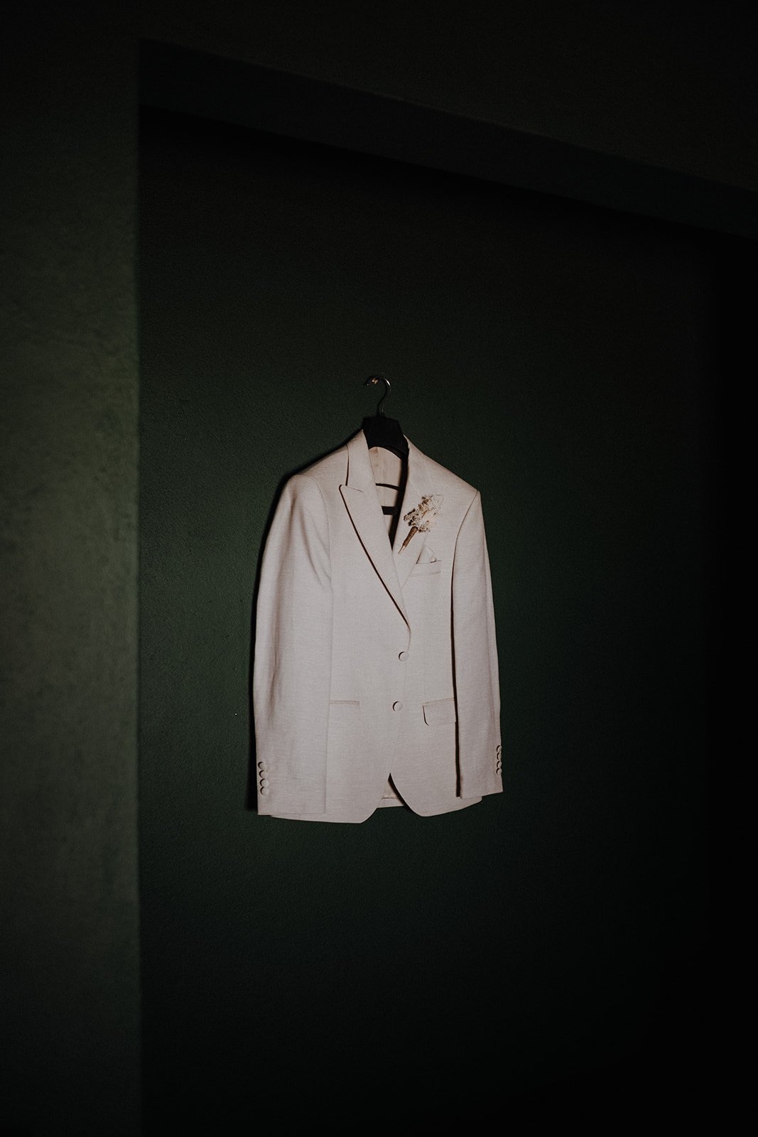 A white suit jacket hanging on a black hanger against a dark green wall.
