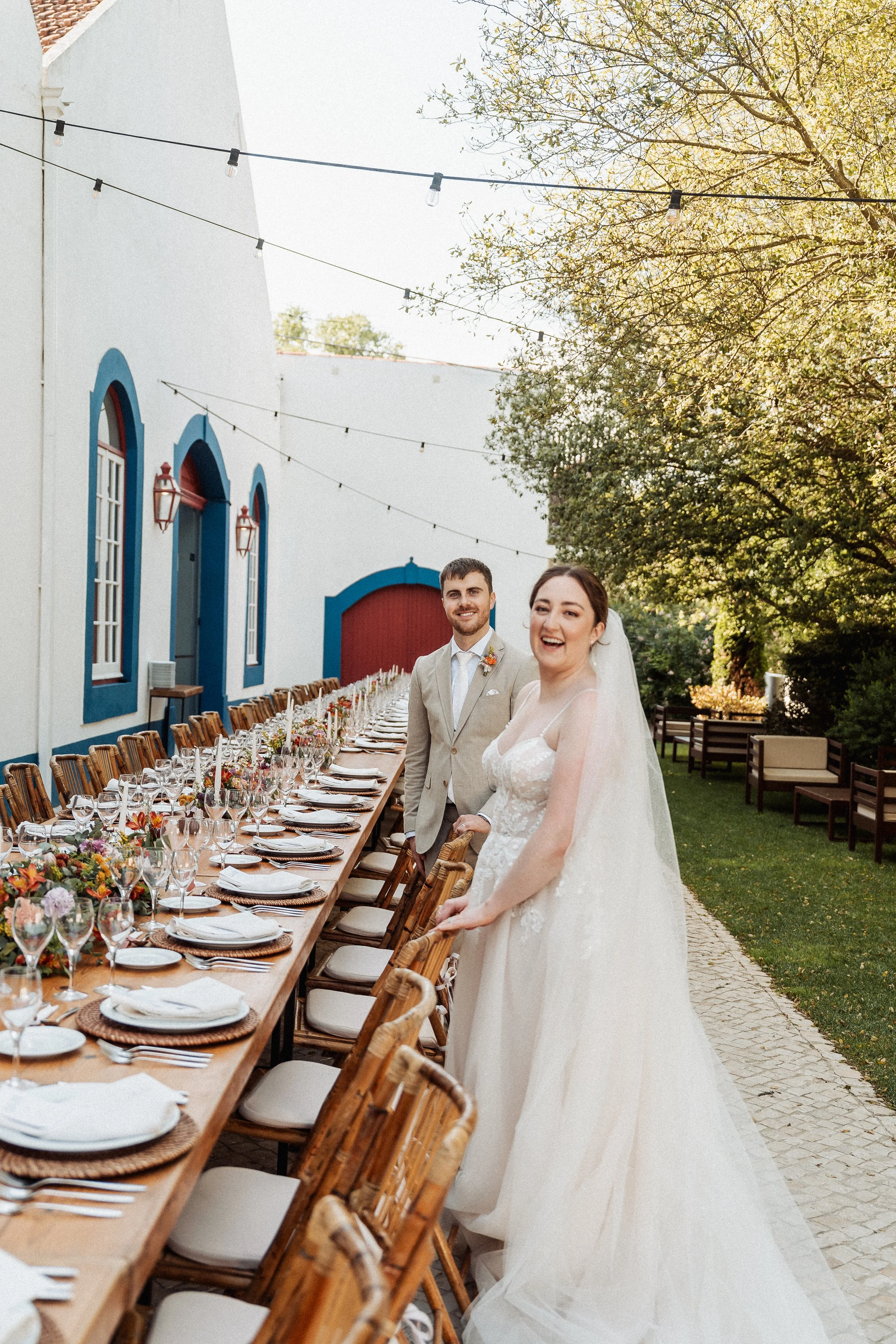 A bride and groom standing at a long outdoor wedding reception table decorated with flowers and tableware, with a white building with blue trim, string lights, and trees in the background. The bride is wearing a white wedding dress and veil, and the groom is dressed in a light suit with a boutonniere. The couple appears happy and smiling.