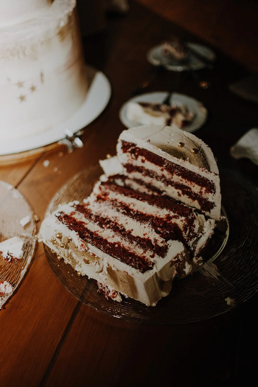 Slice of red velvet cake with cream cheese frosting on a glass plate, set on a wooden table near a white tiered cake.