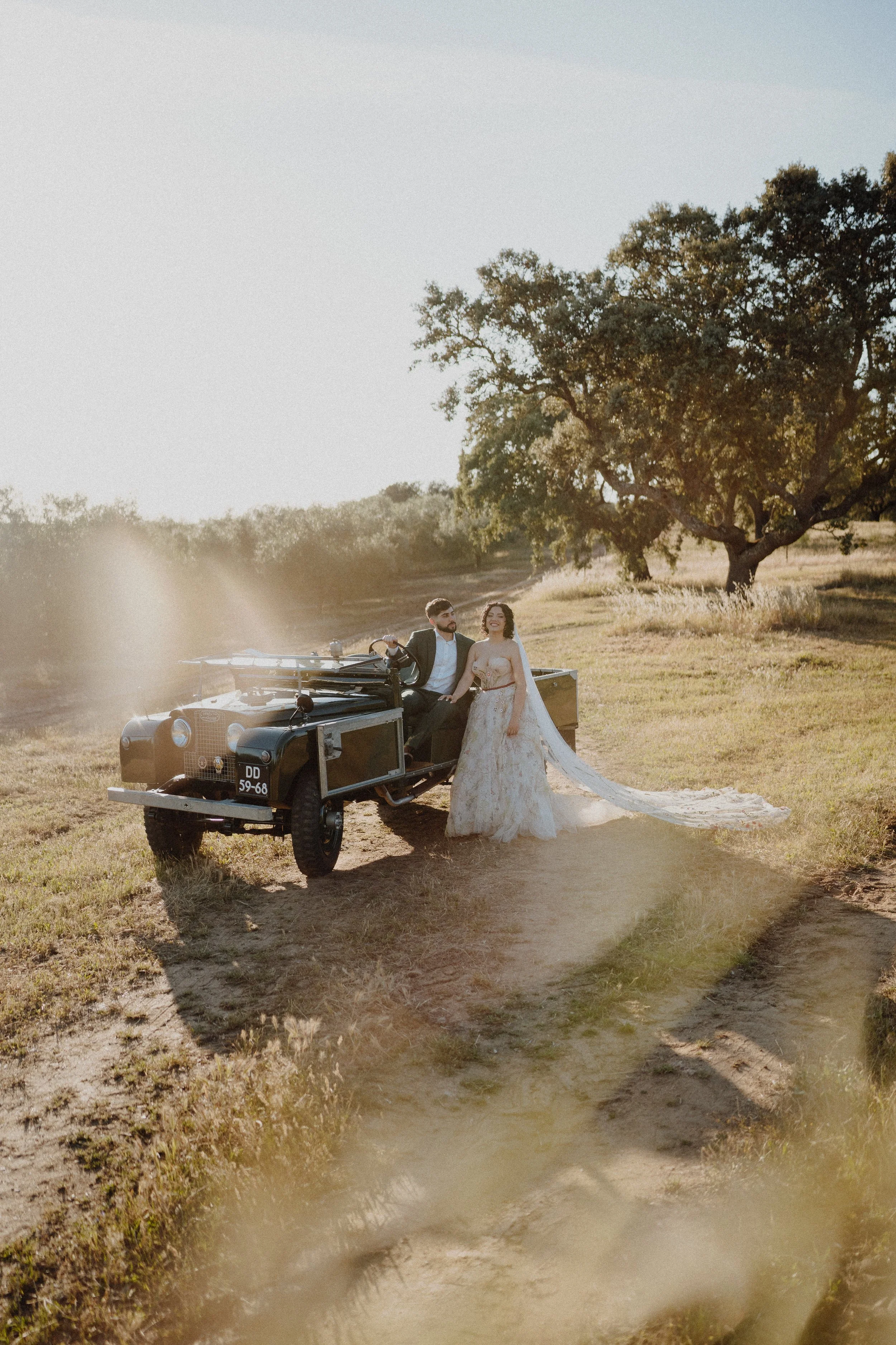 A man and a woman in wedding attire sitting on the hood of an antique black car in a grassy field with trees, during sunset.