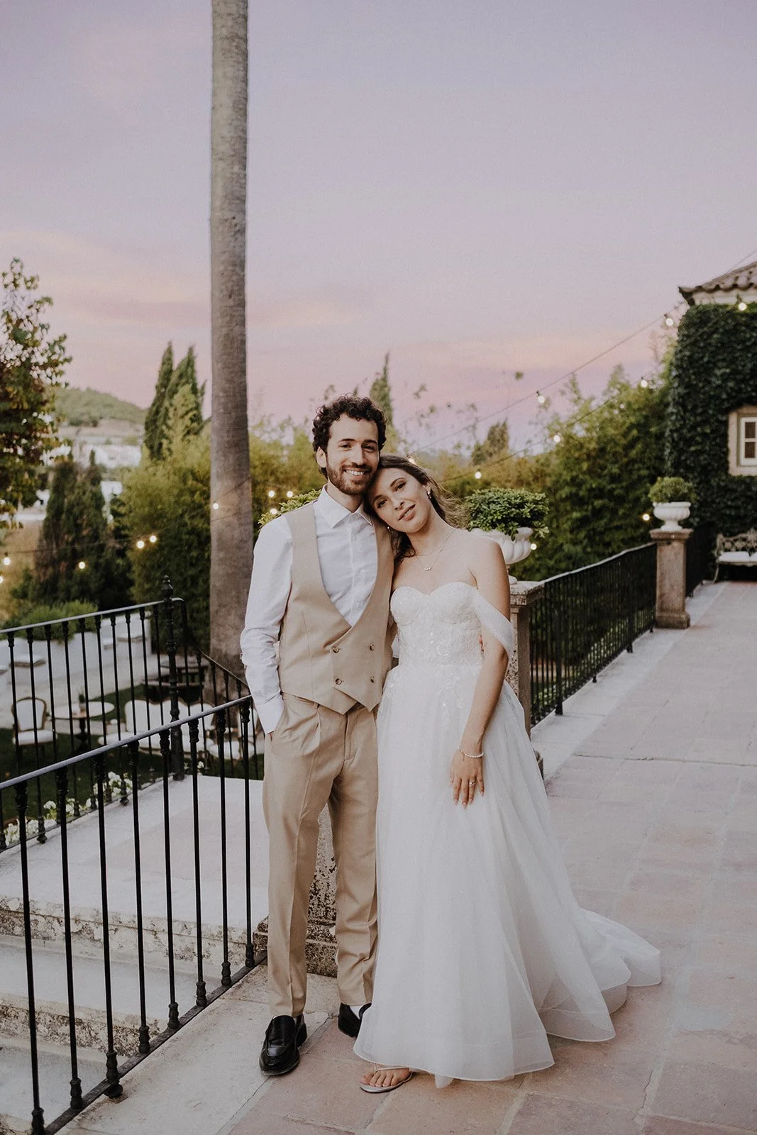 Bride and groom standing together outdoors during sunset, smiling, with a tree and string lights in the background.
