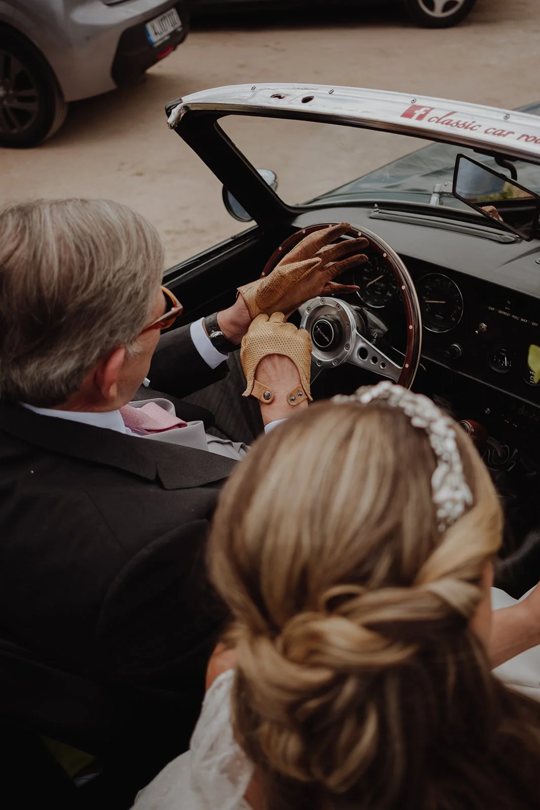 A man and woman dressed in wedding attire sitting in a vintage black convertible car, with the man driving and the woman in the passenger seat, outside in a dusty area.