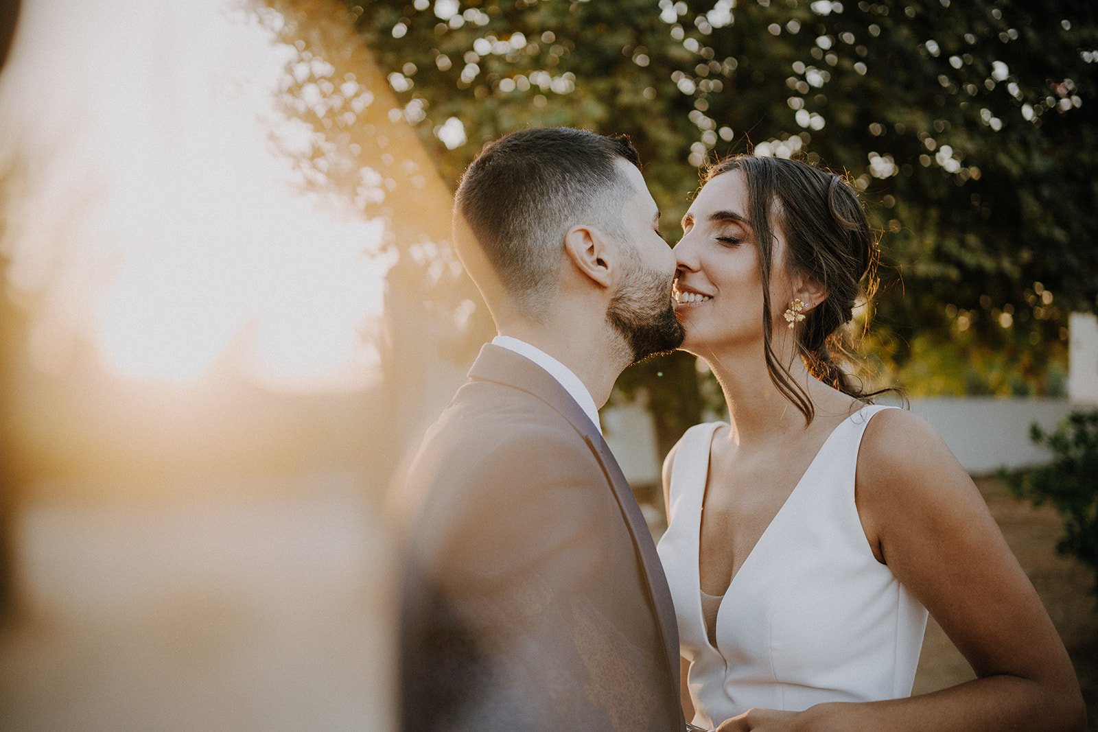 A newlywed couple kissing outdoors at sunset, with trees and a white fence in the background.