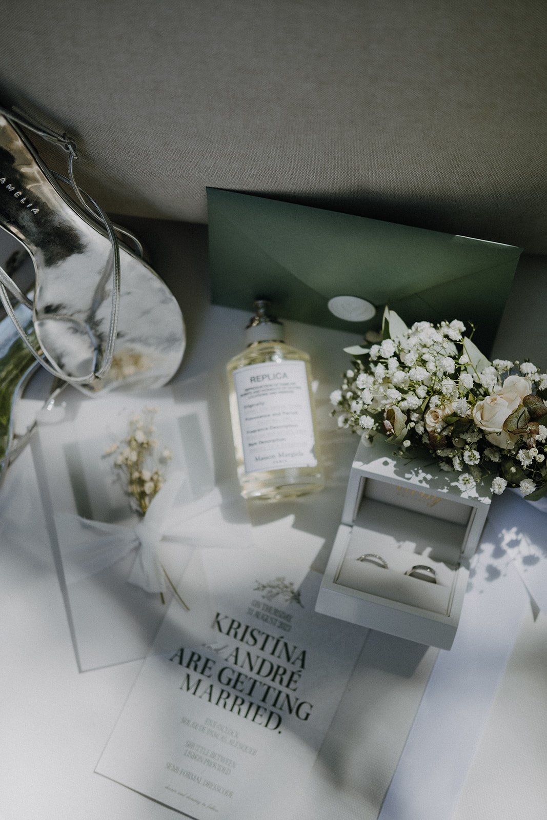 Wedding invitation, wedding rings in a white box, bouquet of white flowers, perfume bottle, and a pair of glasses on a table.