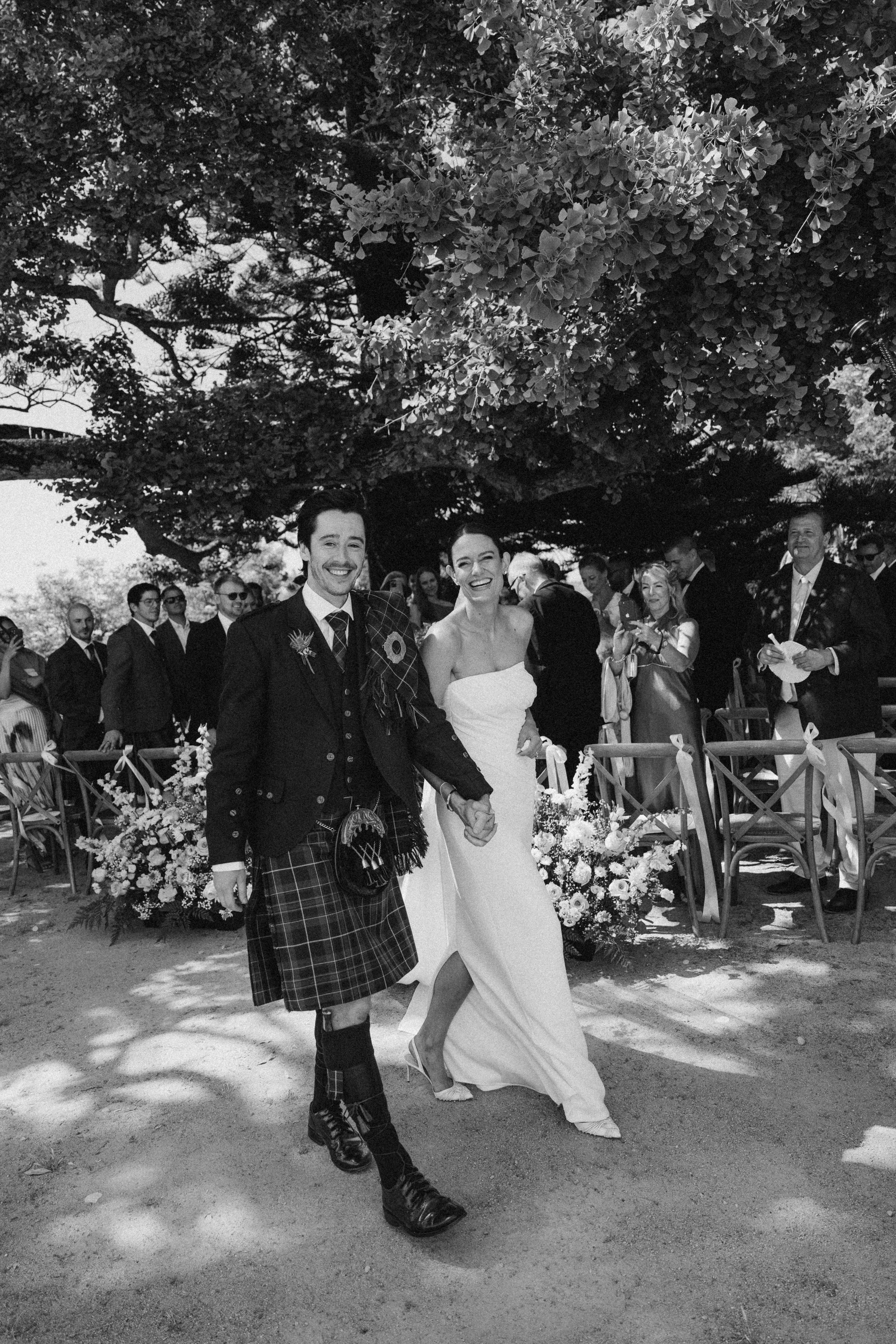 Black and white photo of a bride and groom holding hands, walking down the aisle at their wedding ceremony outdoors under a large tree, surrounded by guests and floral arrangements. Quinta São Thiago.