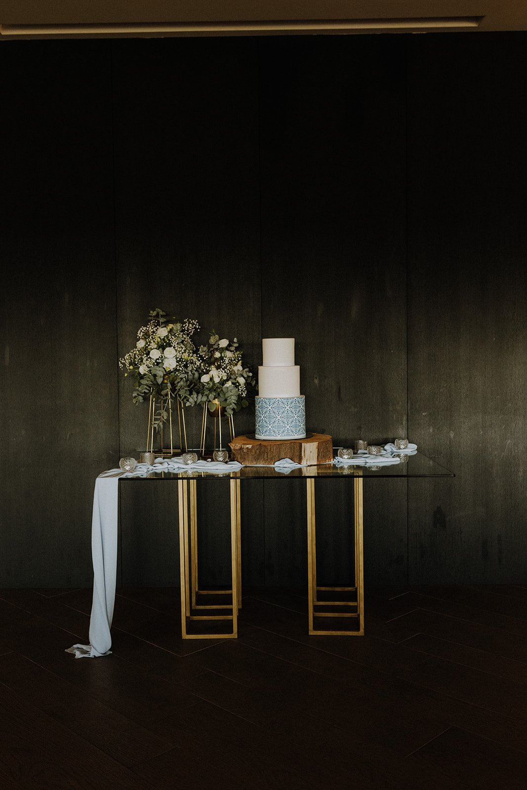 Elegant three-tier wedding cake with a blue patterned band on the middle tier, placed on a wooden slab, decorated with a flower arrangement, on a glass table with gold legs.