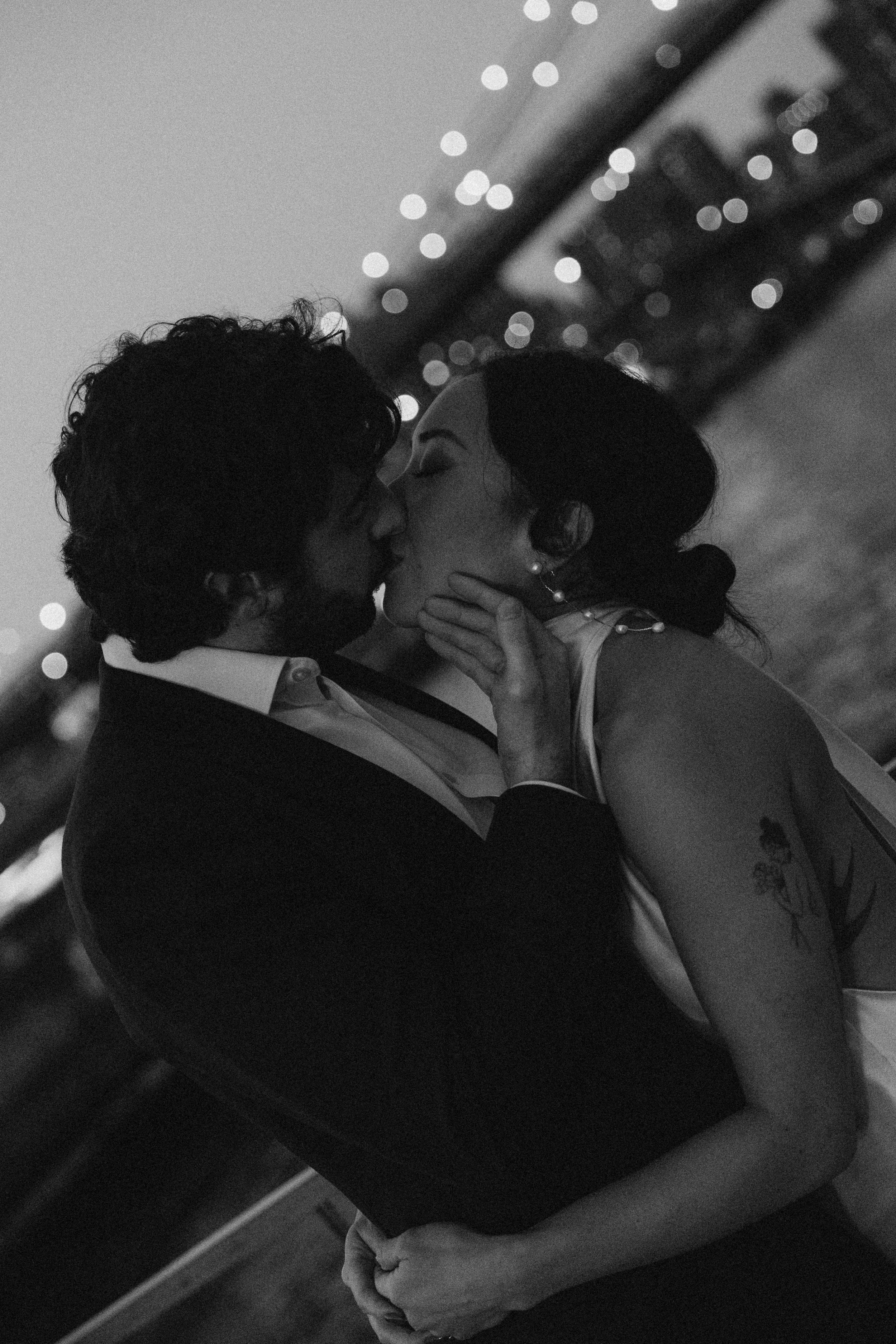 A black and white photograph of a couple kissing closely, with the woman's hand resting on the man's shoulder. The scene is lit with string lights in the background, creating a romantic atmosphere.