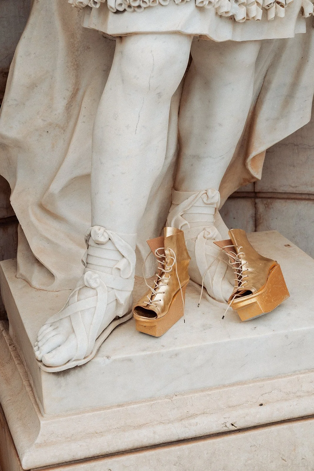 Close-up of stone statue's feet wearing ancient sandals, with modern gold platform lace-up shoes placed in front. Vandelli Gardens, Lisbon.