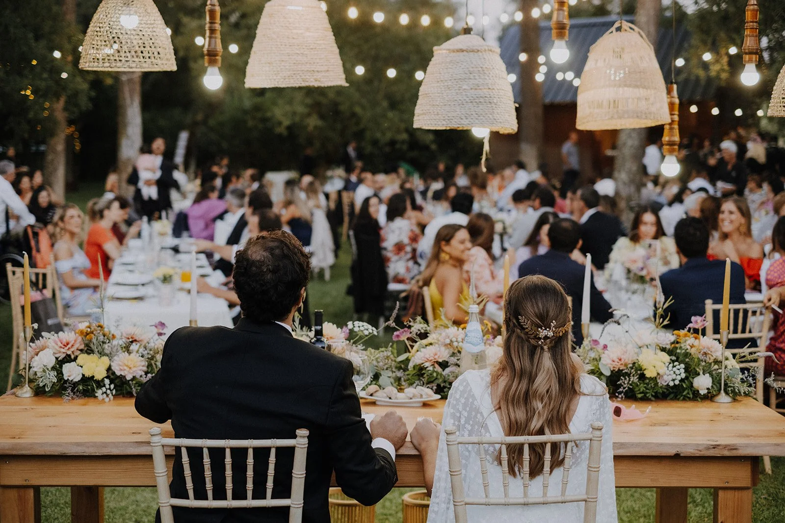 Wedding reception outdoors at night with string lights, a large gathered crowd, and a bride and groom sitting at a wooden head table decorated with flowers