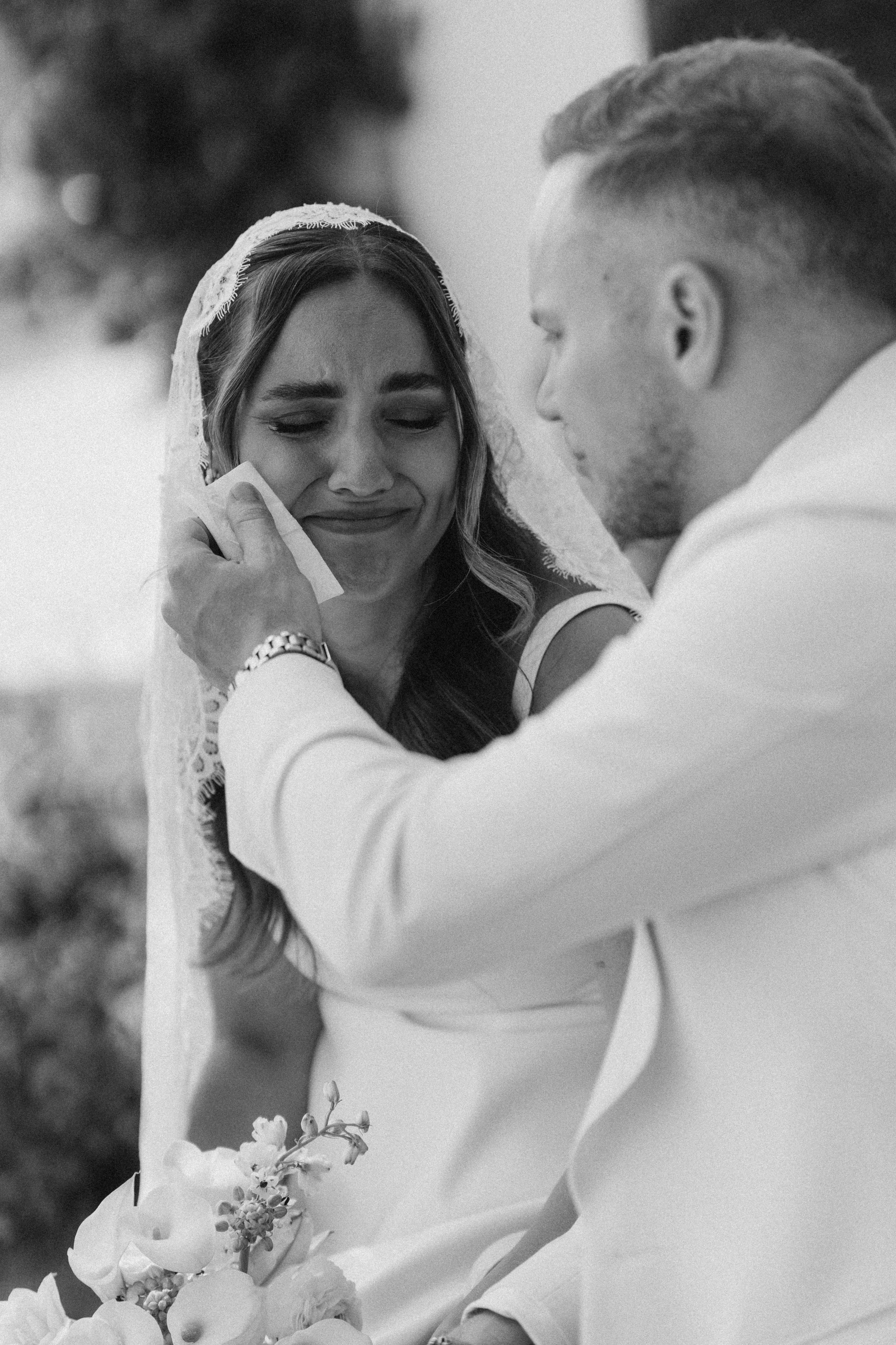 A black-and-white photo of a woman in a wedding dress with a lace veil, tears in her eyes, being comforted by a man who is gently holding her face and kissing her forehead. The woman holds a bouquet of flowers.