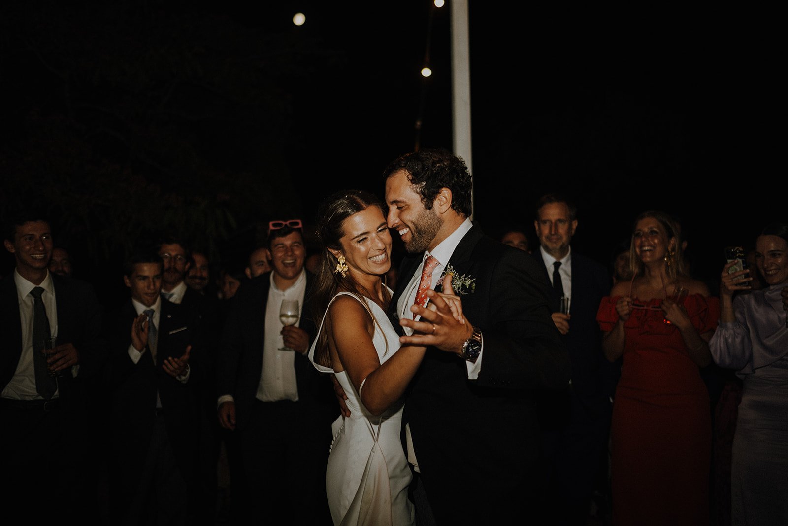 A bride and groom share a dance at their wedding reception, surrounded by smiling guests in formal attire.