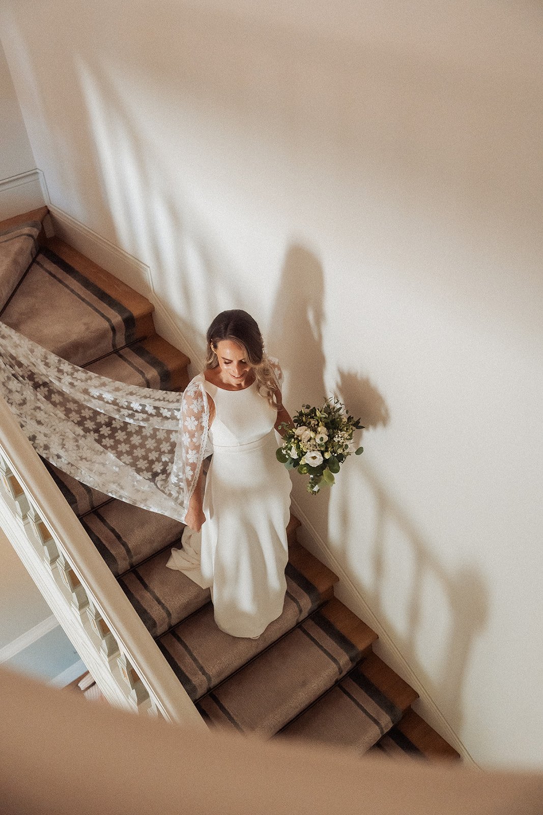 Bride ascending a carpeted staircase, holding a bouquet of white and green flowers, with a lace veil trailing behind her, wearing a fitted white wedding gown with lace sleeves.