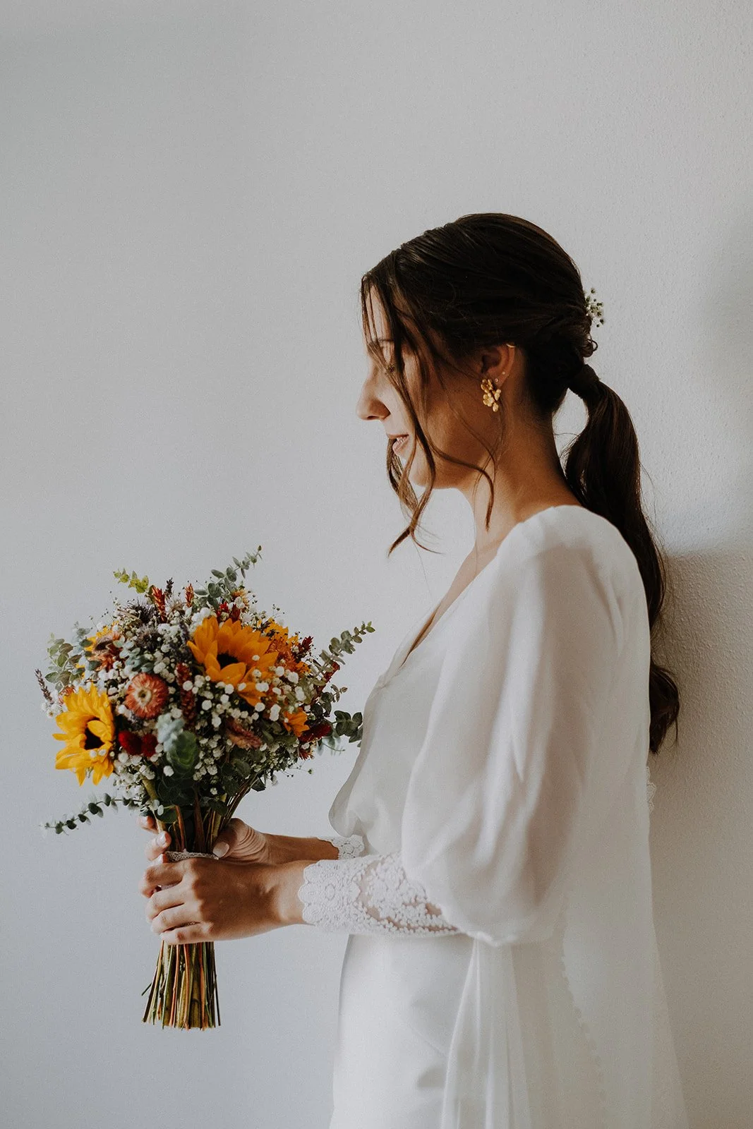 A bride in a white dress holding a bouquet of sunflowers, daisies, and greenery, standing sideways against a plain wall, with her hair in a ponytail and gold earrings.