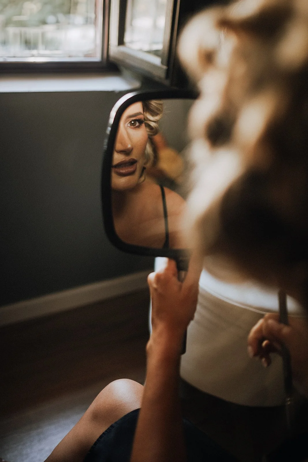 Bride holding a small mirror, looking at their reflection which shows their face, while sitting on the floor near a window.