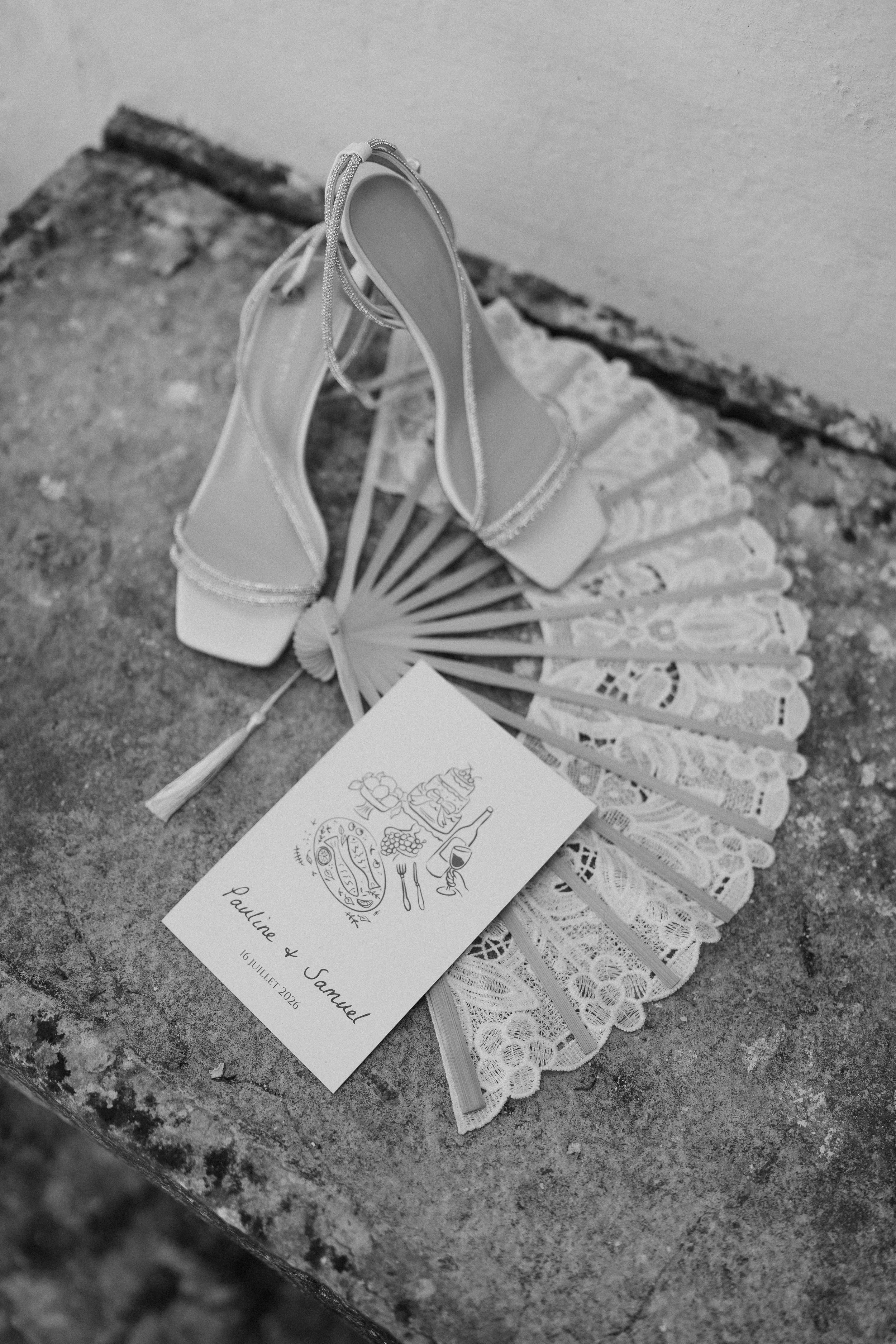 A wedding invitation card resting on a lace fan, placed on a rough textured stone surface. The invitation shows an illustration of a cake, wine glasses, and food items, with the names Paulina and Samuel and the date 16 July 2026 written on it. Two hi
