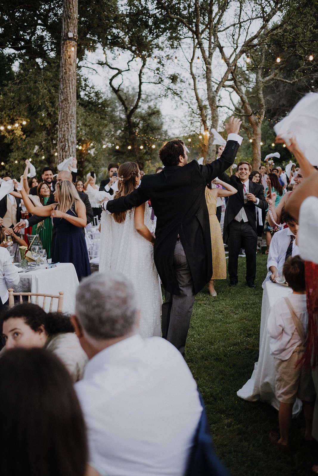 Guests dancing and celebrating outdoors at a wedding reception with string lights, surrounded by trees and tables.