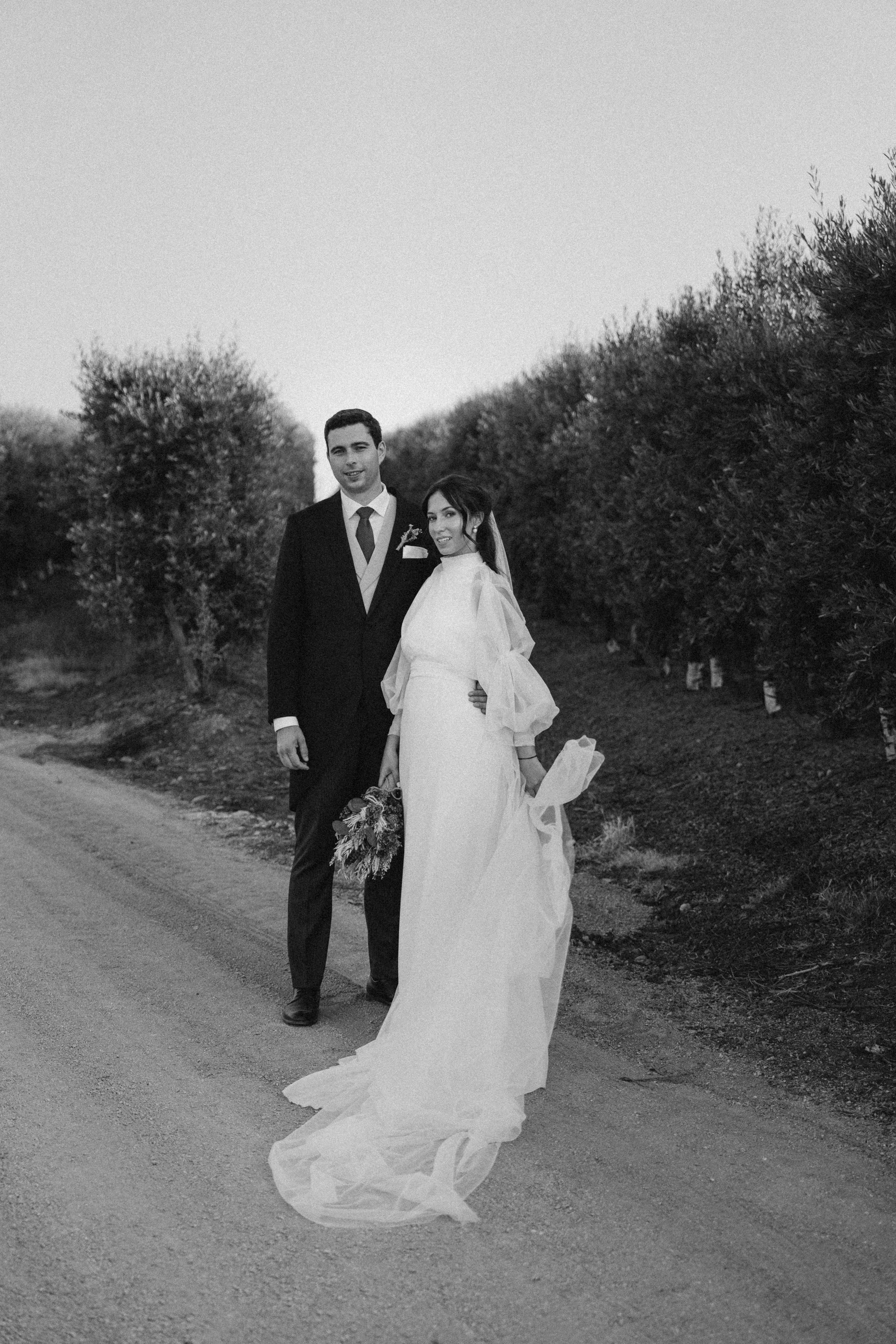 A black and white photo of a bride and groom standing on a dirt road with bushes on either side. The groom wears a dark suit with a tie, and the bride wears a long white gown with puffed sleeves, holding a bouquet of flowers. They are smiling and fac