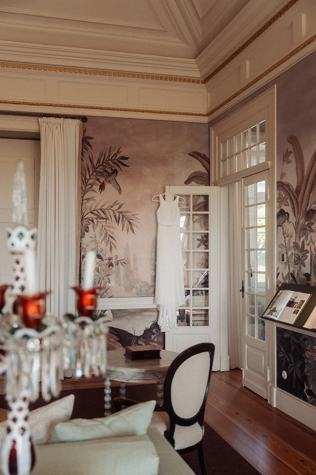 A white wedding dress hanging on a door in front of a painted mural of tropical plants in a room with ornate ceiling molding, dark wooden flooring, and glass-paneled doors.