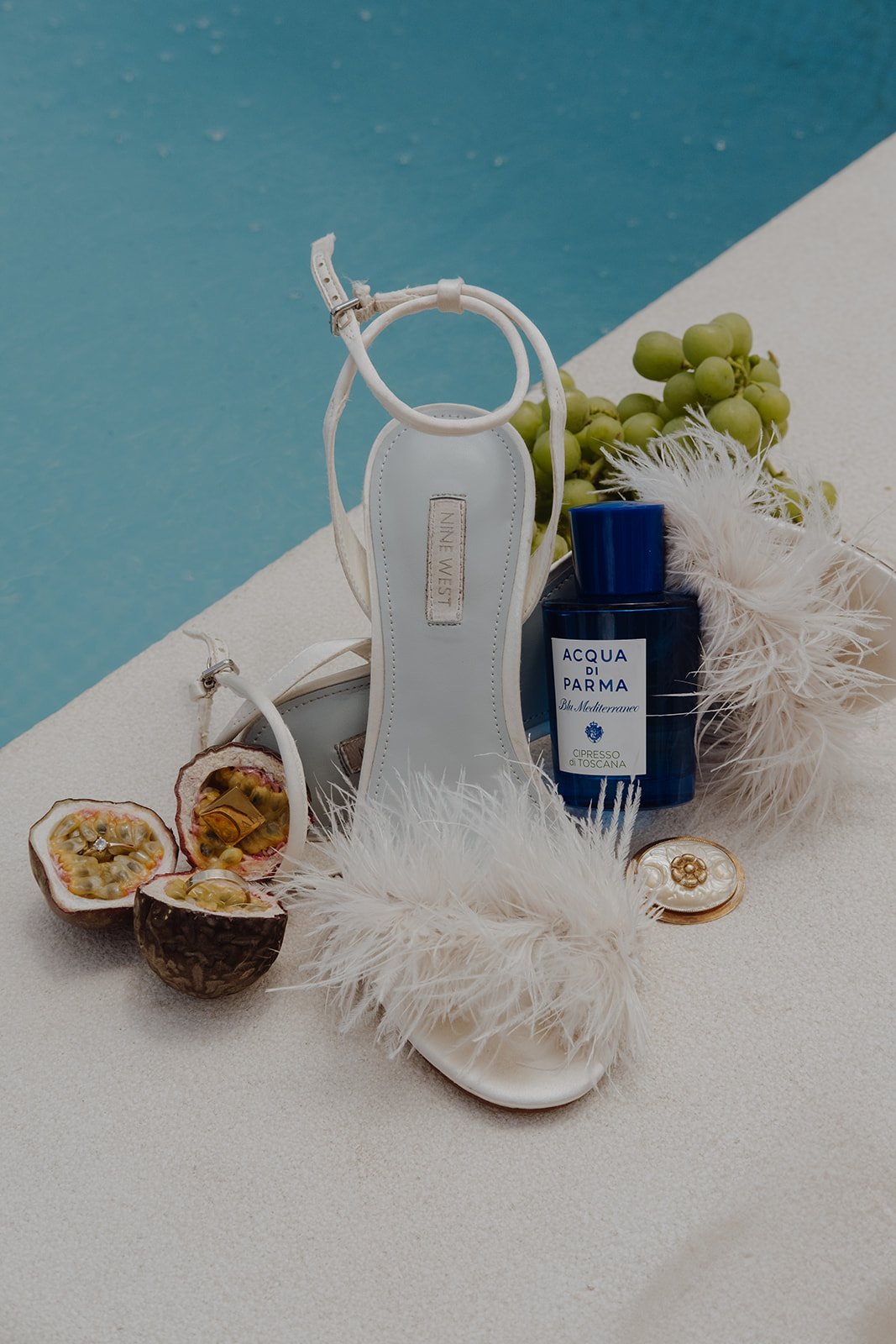 Wedding deatails. perfume bottle labeled 'Acqua di Parma Blu Mediterraneo,' white feather accessory, white strap, gold ring with decorative stone, small bottle, and a white-and-blue background.