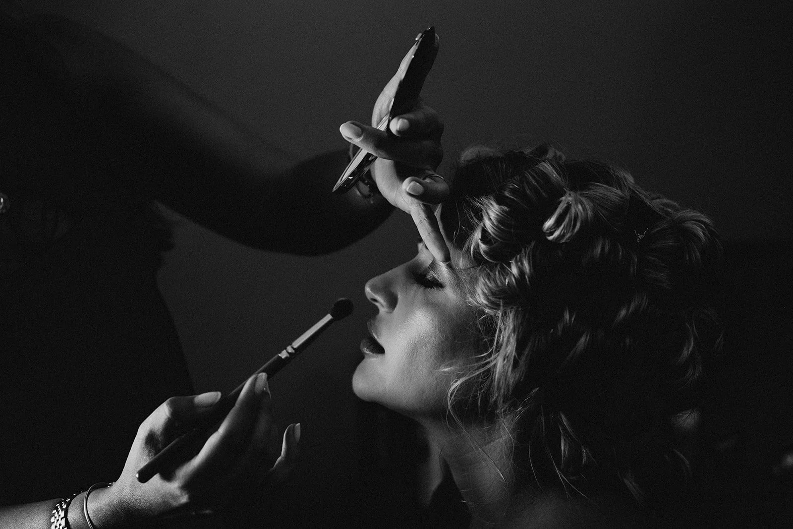 Black and white photo of a woman with curled hair having makeup applied, with her eyes closed and lips slightly parted.