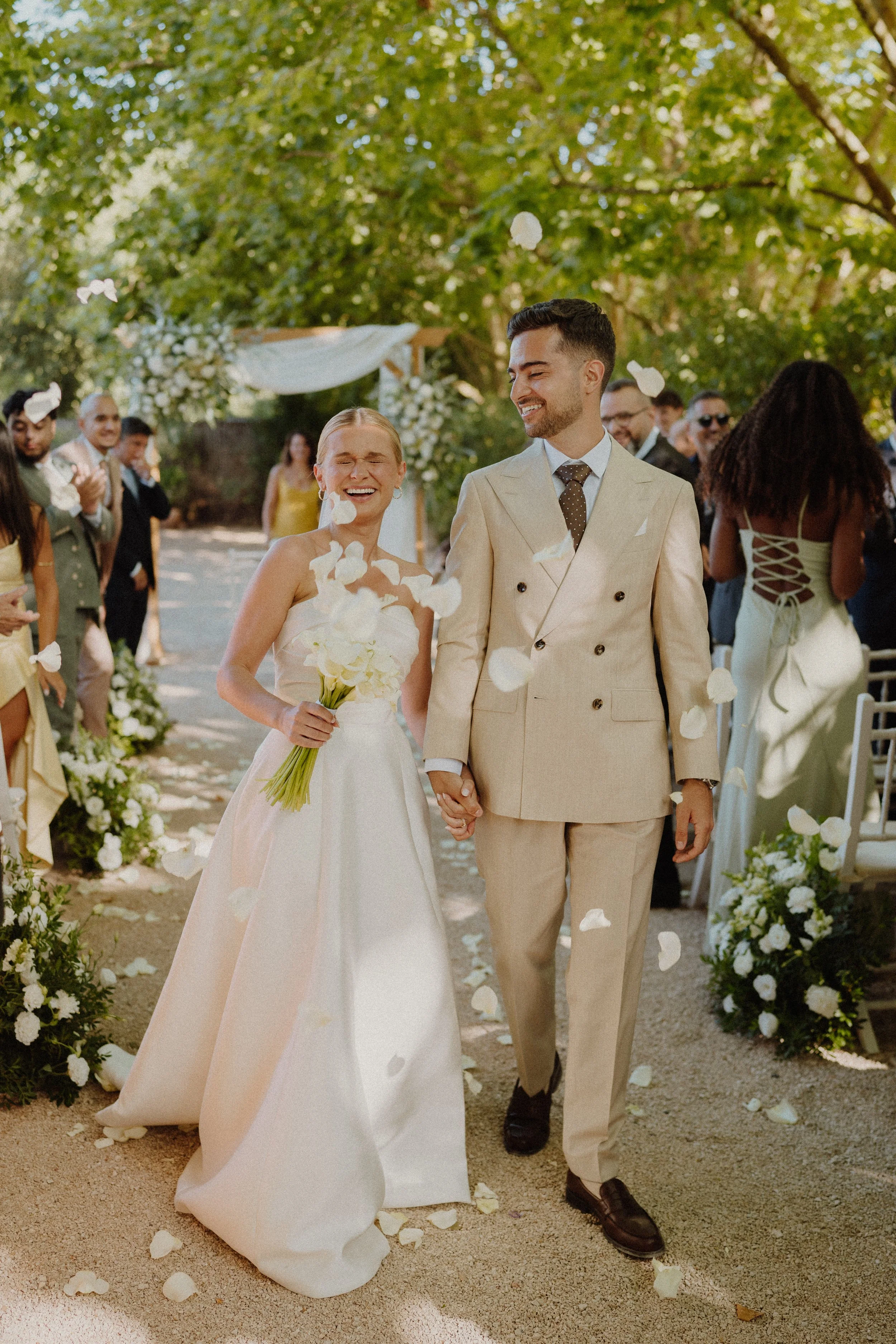 A newlywed couple walking hand in hand down an aisle decorated with white flowers, surrounded by guests at an outdoor wedding ceremony with green trees in the background, as white flower petals fall around them.