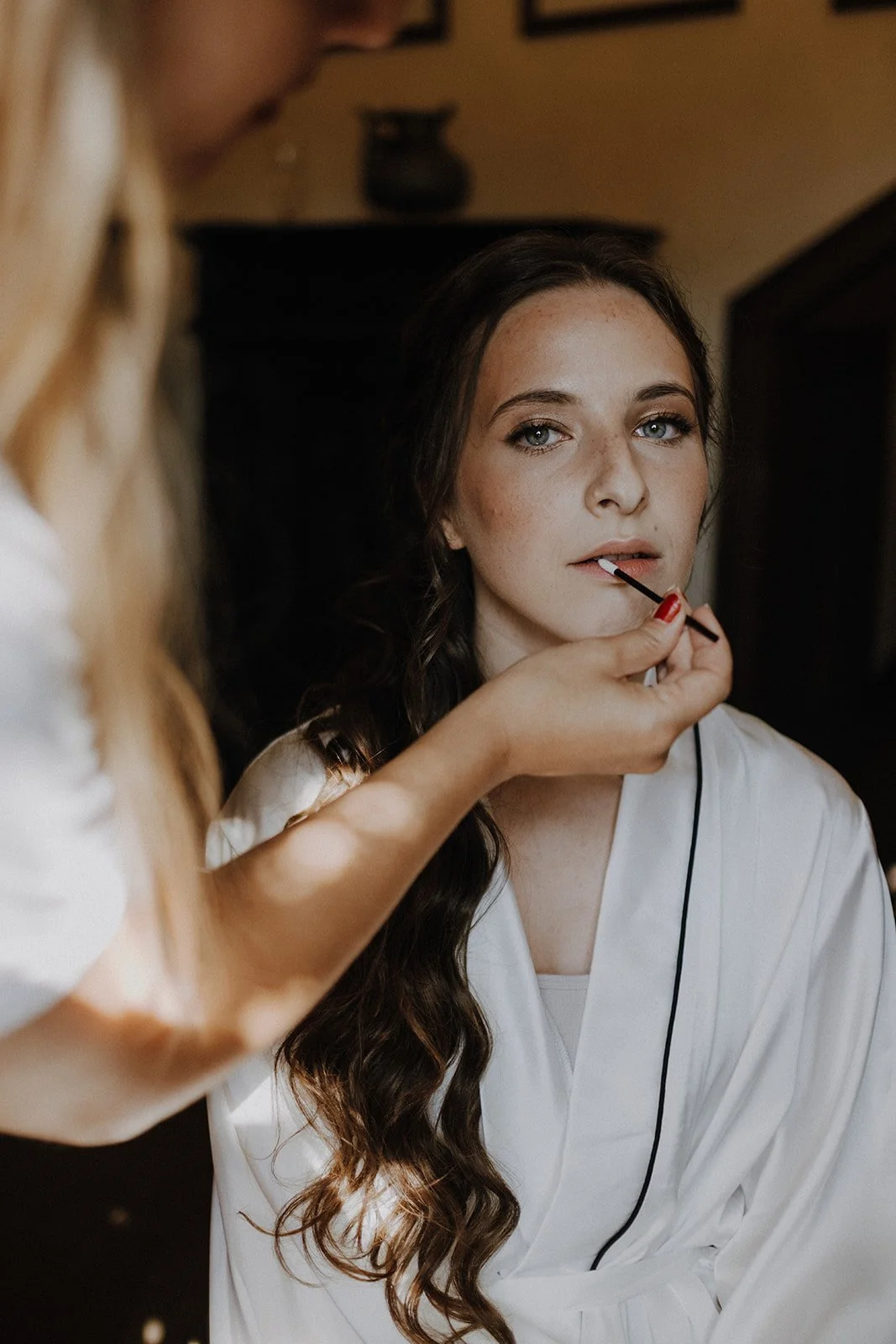 Bride getting her makeup done by a makeup artist, who is applying lipstick with a brush.