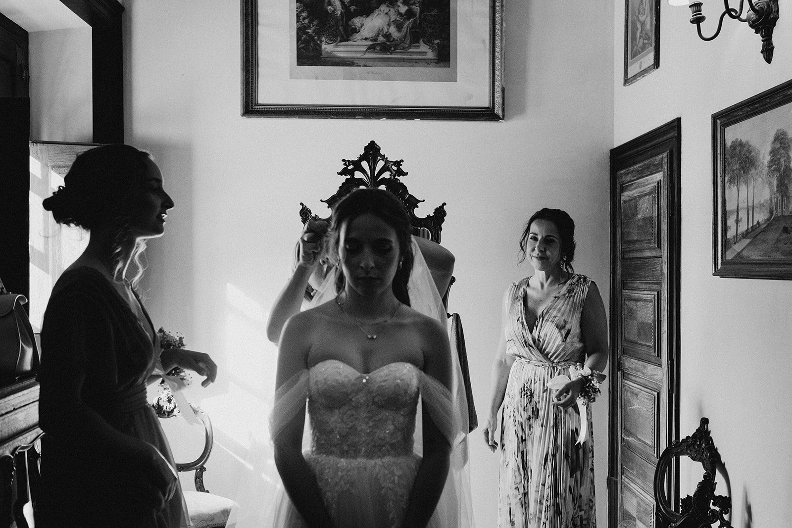 A bride in a strapless wedding dress preparing with her bridal party in a living room with framed paintings on the wall.