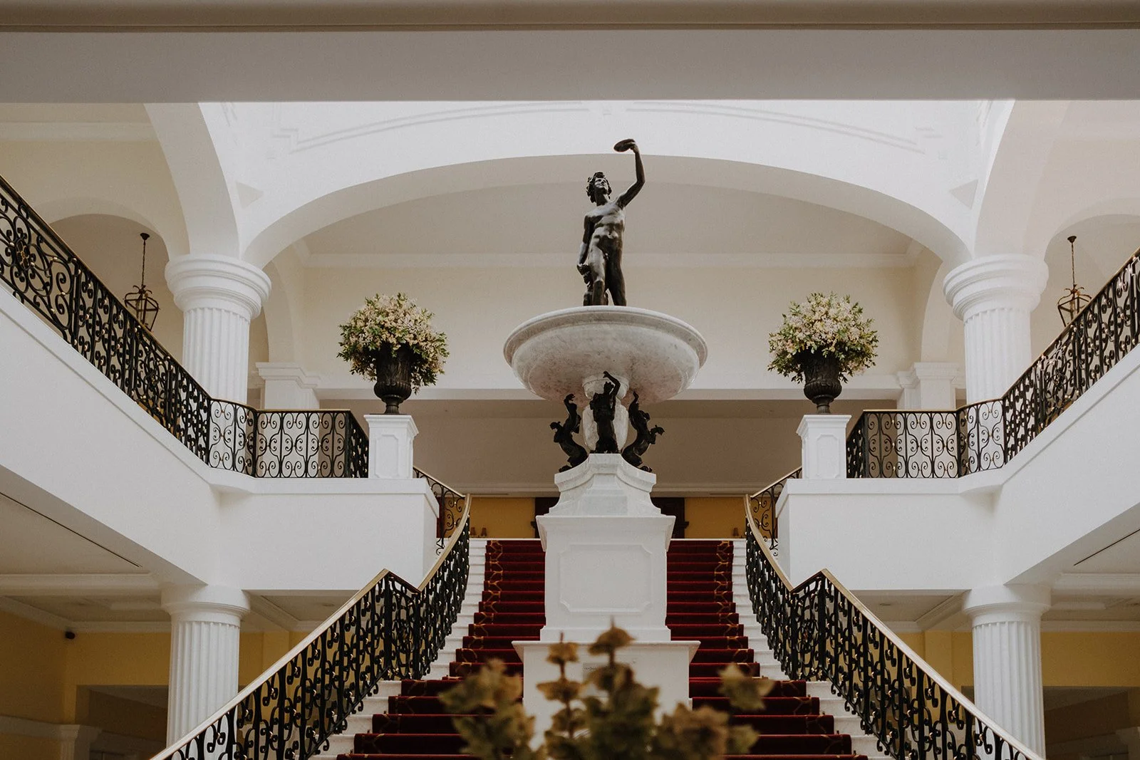Elegant hotel lobby with a central staircase, ornate black wrought iron railings, a statuesque sculpture at the top of a fountain, and floral arrangements in large vases.